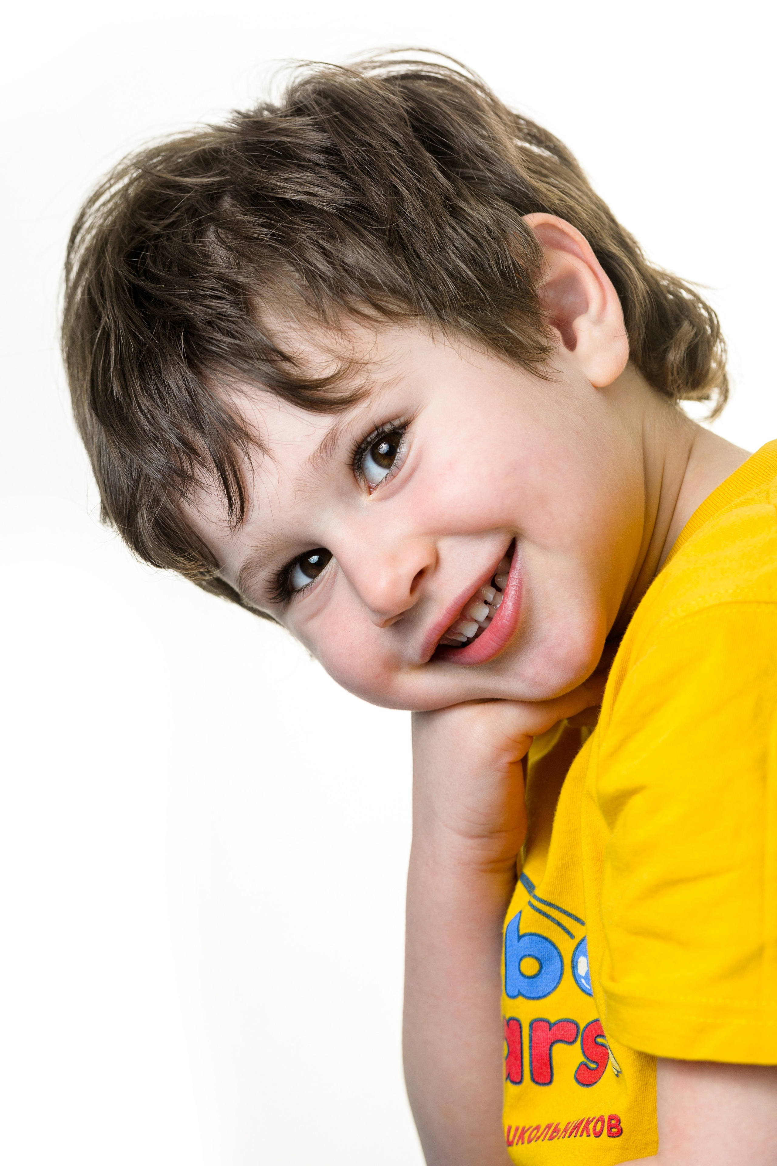 Мальчик в жёлтой футболке выглядывает сбоку и улыбается в студийном кадре на белом фоне.