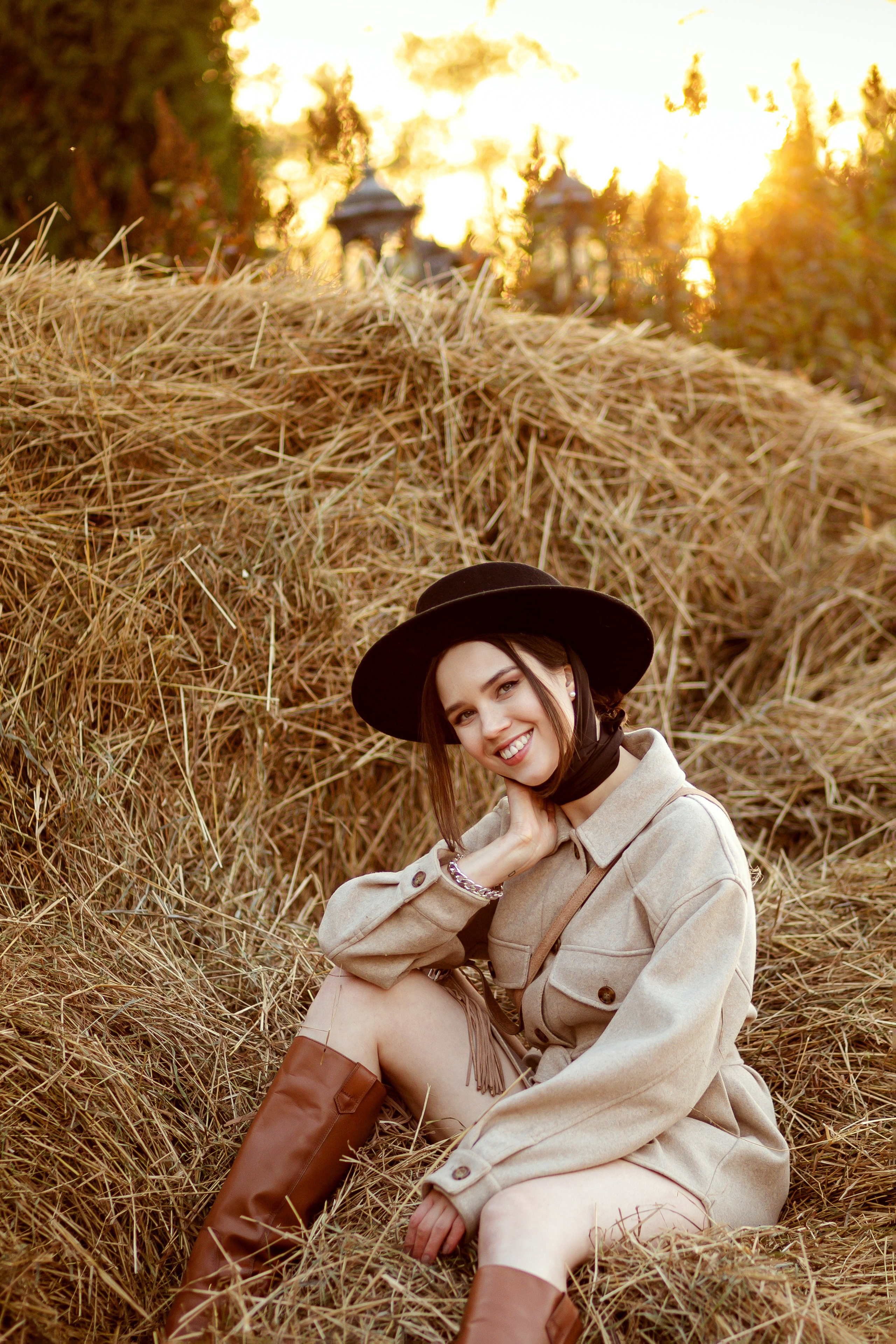 Улыбающаяся девушка сидит на стоге сена, за стогом садится солнышко.