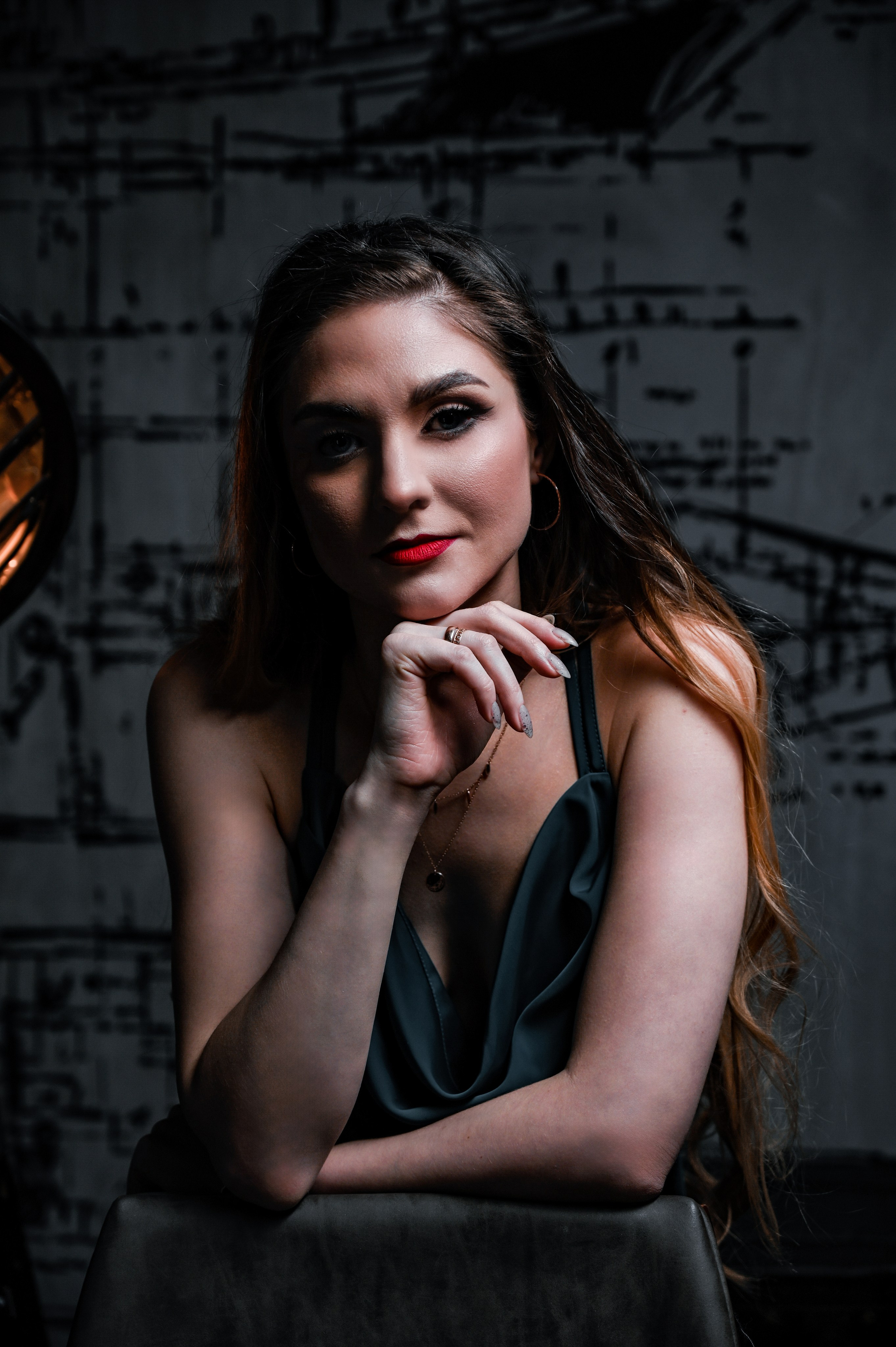 Молодая девушка сидит в темной комнате, создавая атмосферу загадочности и элегантности. Она одета в зелёное платье с открытыми плечами и имеет длинные волосы, которые свободно падают по плечам. Ее взгляд направлен прямо на камеру, демонстрируя уверенность и сосредоточенность. На лице у нее акцентный макияж с ярко-красными губами, который контрастирует с тёмным фоном. Рука поддерживает подбородок, добавляя игривость и задумчивость к образу. В фоне видны графические элементы на стене, а слева от модели расположено освещение, создающее мягкое свечение и акцентирующее внимание на её лице. Фотография передает чувственную и стильную атмосферу.