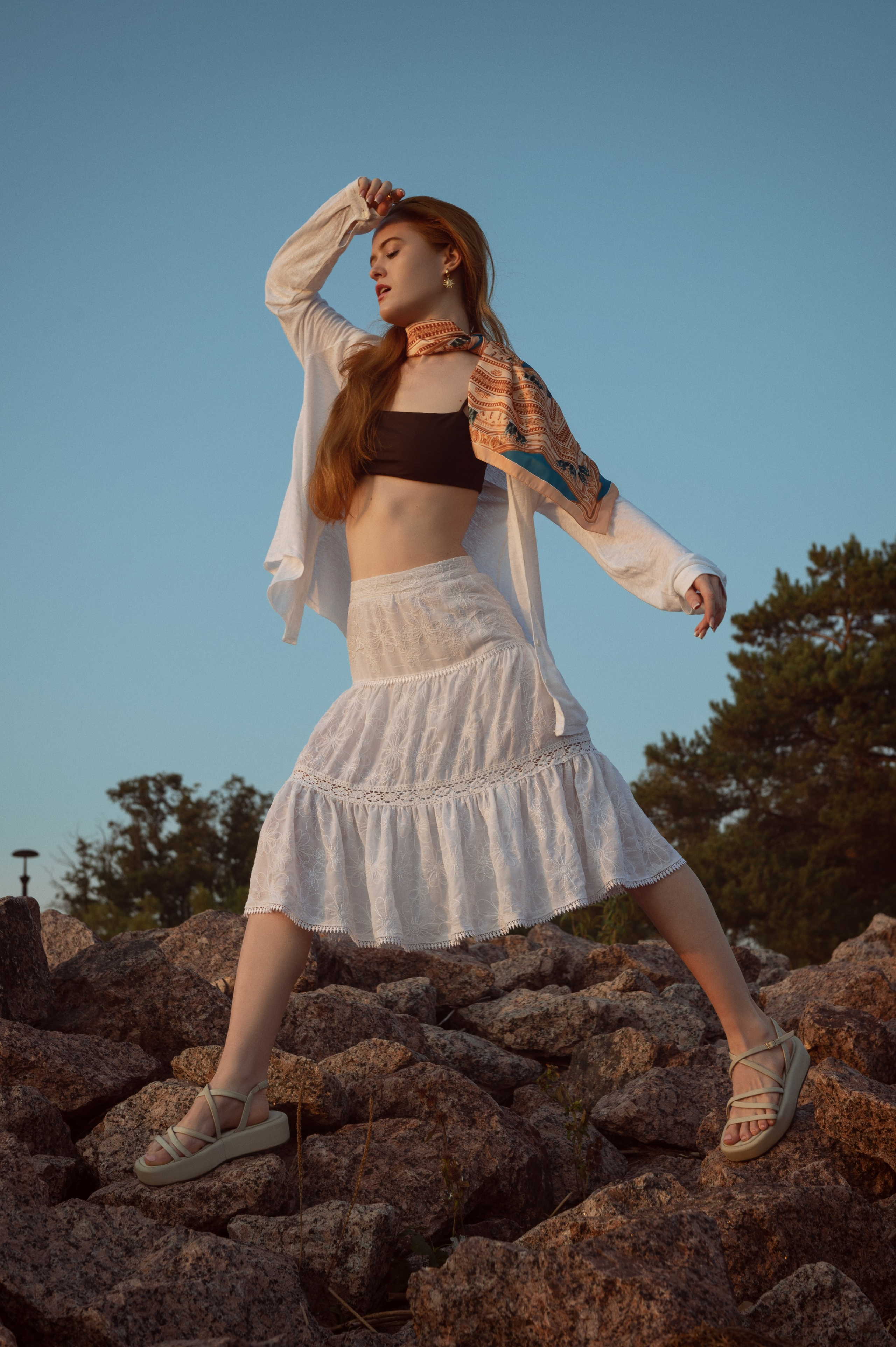 Рыжеволосая девушка в белой юбке-миди, белой рубашке и топе на фоне голубого неба и камней. На шее у нее развевается яркий желтый платок.