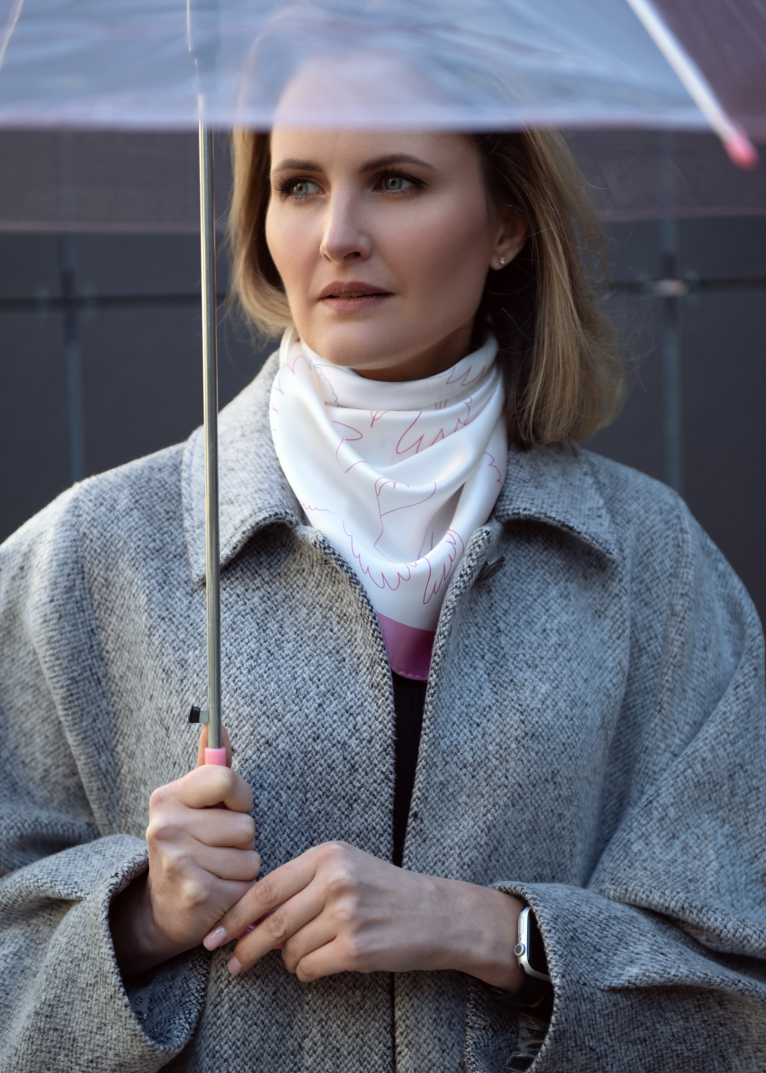 Красивая женщина с прозрачным зонтом в сером пальто и белом шелковом платке стоит на фоне стены и смотрит вдаль. Съемка с естественным светом. Красивый уличный портрет.