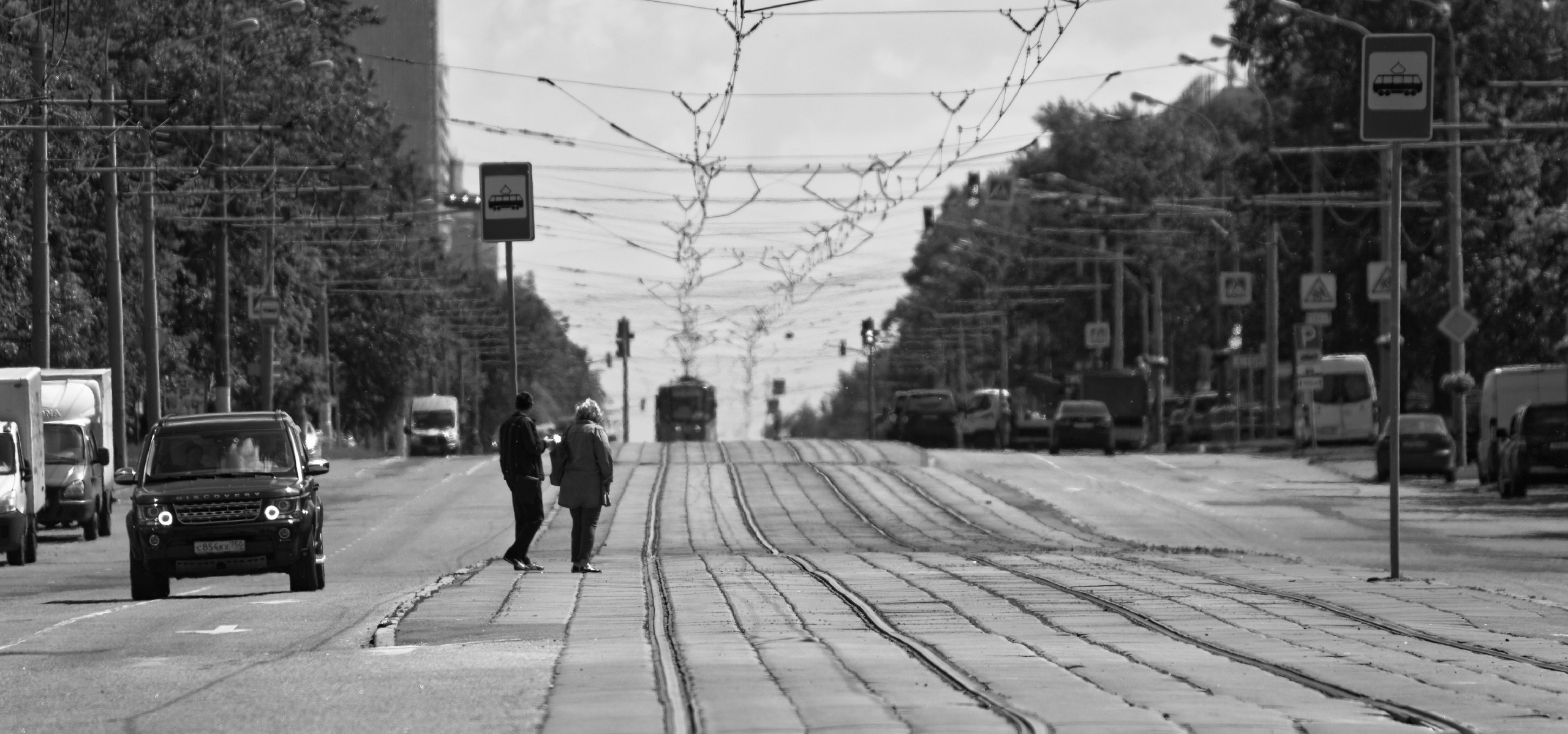 Два человека идут по трамвайным путям под переплетением контактных проводов, трамвай вдали, черно-белое фото городской инфраструктуры