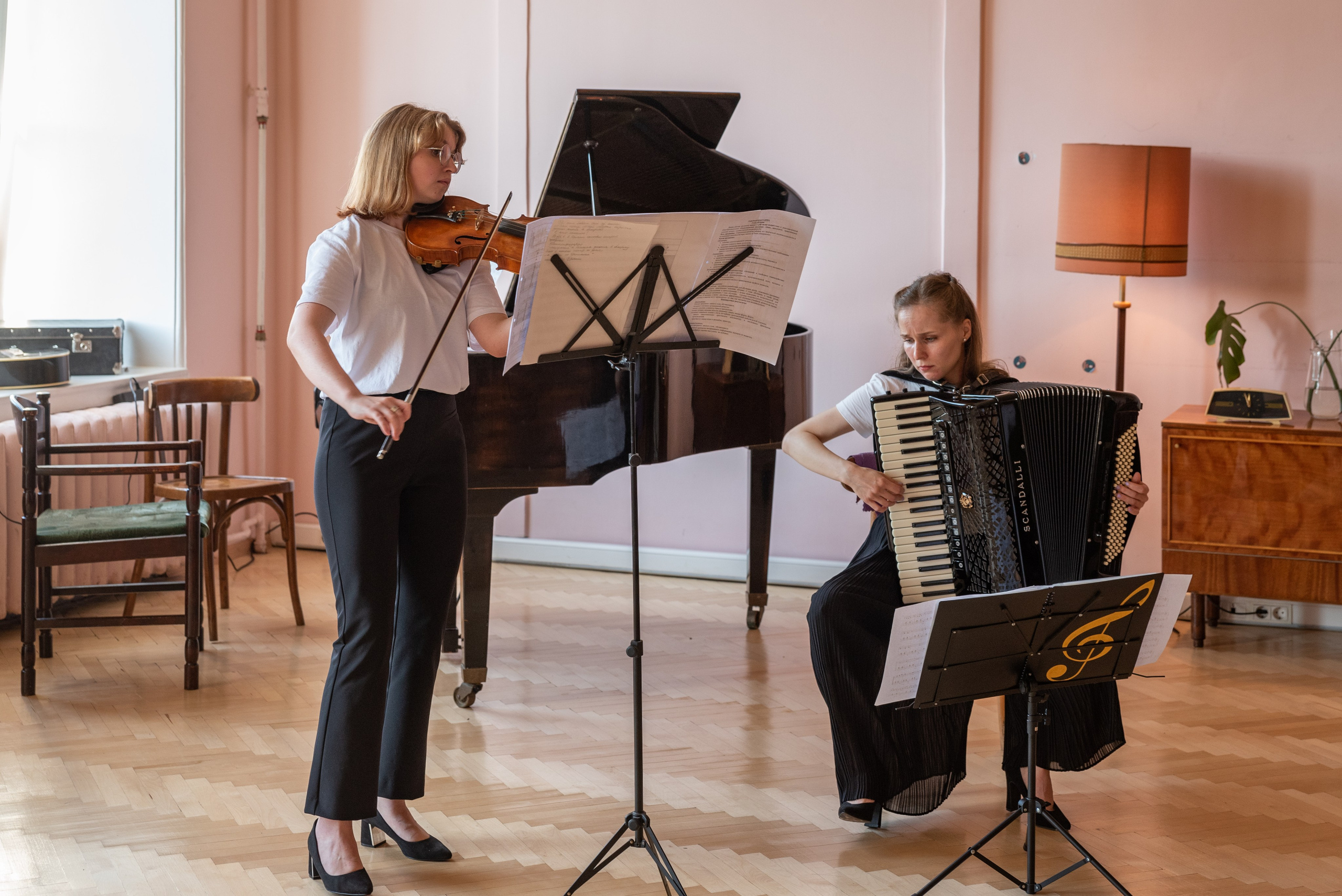 Две девушки играют на аккордеоне и скрипке. Перед ними стойки с нотами, сзади рояль. 