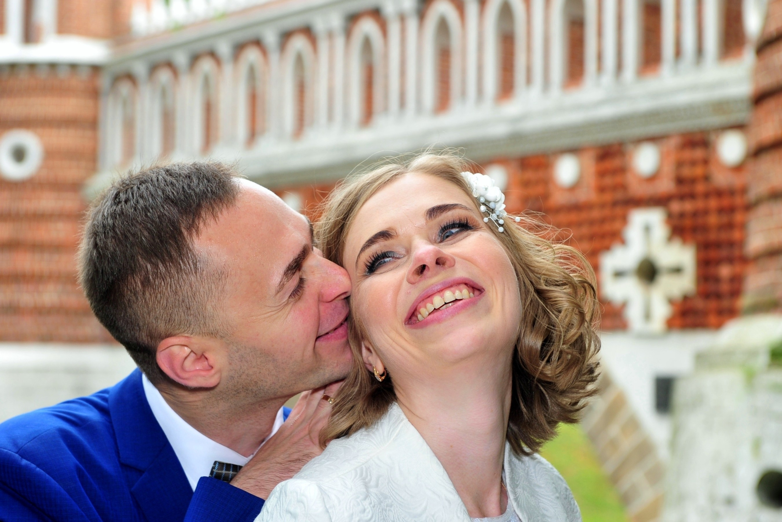 Свадьба в Царицыно хмурым прохладным летним днём, жених целует невесту в щёку на фоне моста из красного кирпича и белого камня