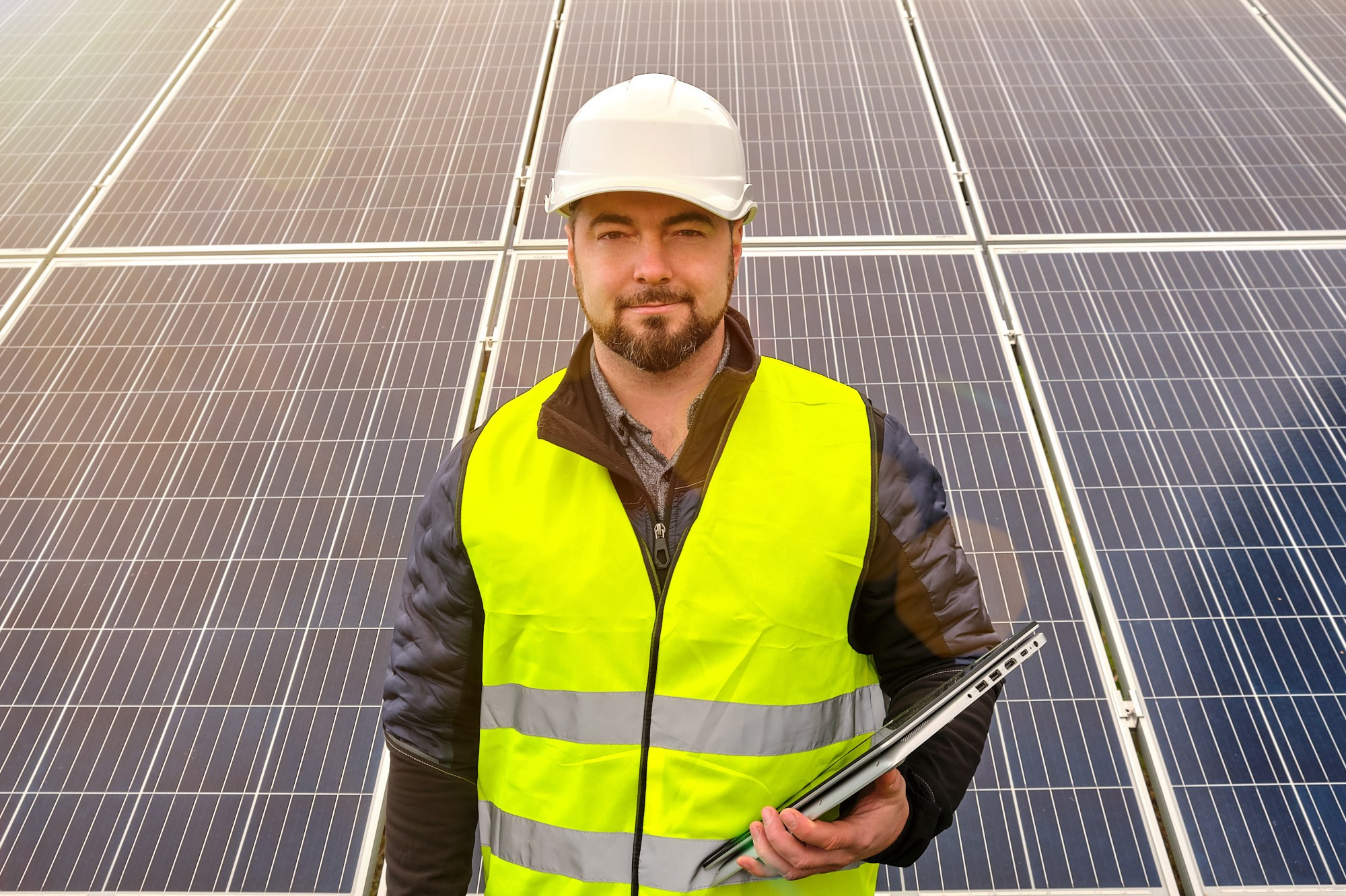 Коммерческая фотосъёмка инженера на солнечной электростанции: мужчина в защитном жилете и каске проводит инспекцию солнечных панелей. Корпоративный контент для энергетических компаний.