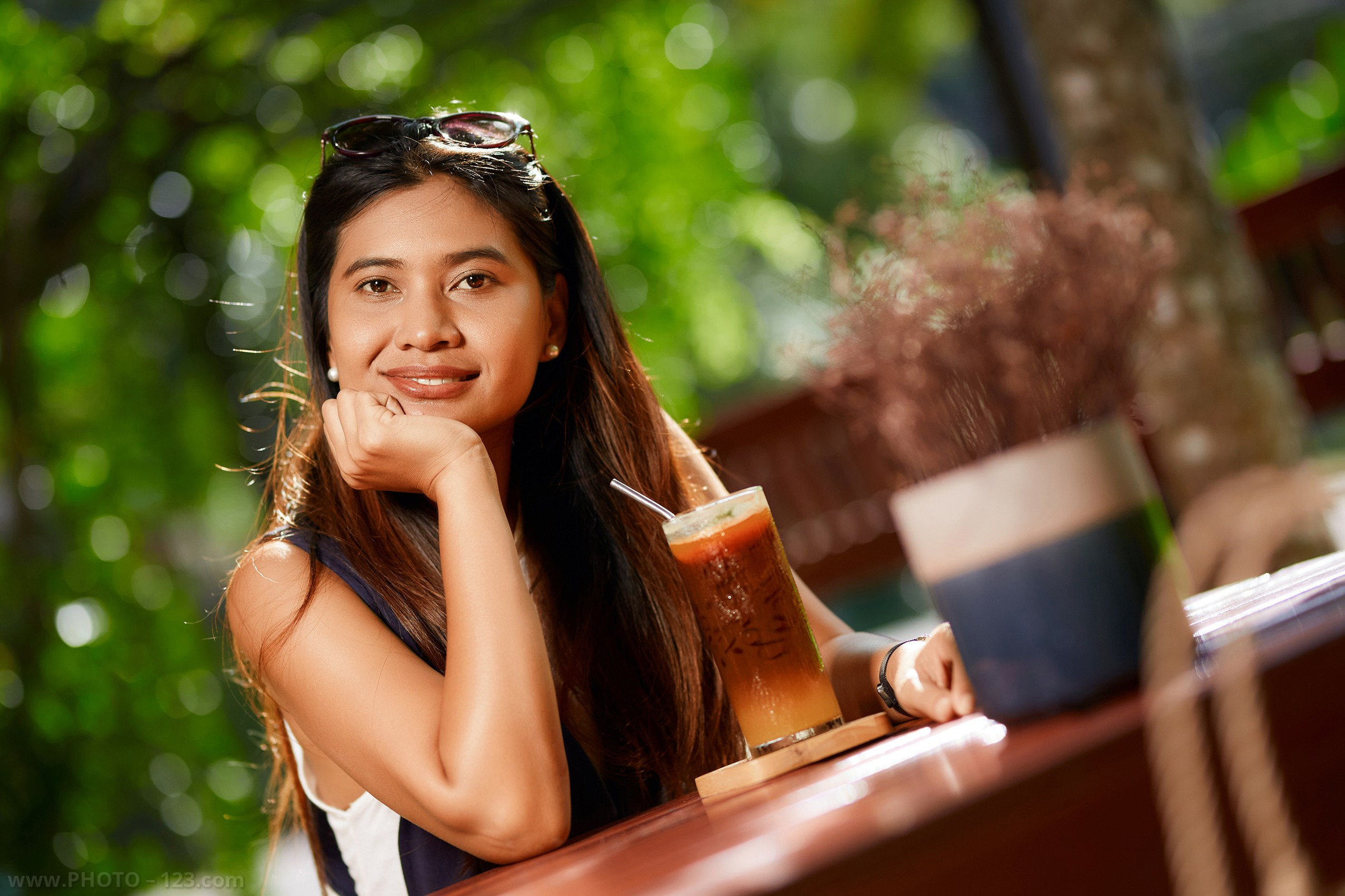Portrait of a woman sitting at an outdoor café table in a green garden, smiling at the camera with a cold drink and soft natural sunlight, relaxed lifestyle atmosphere