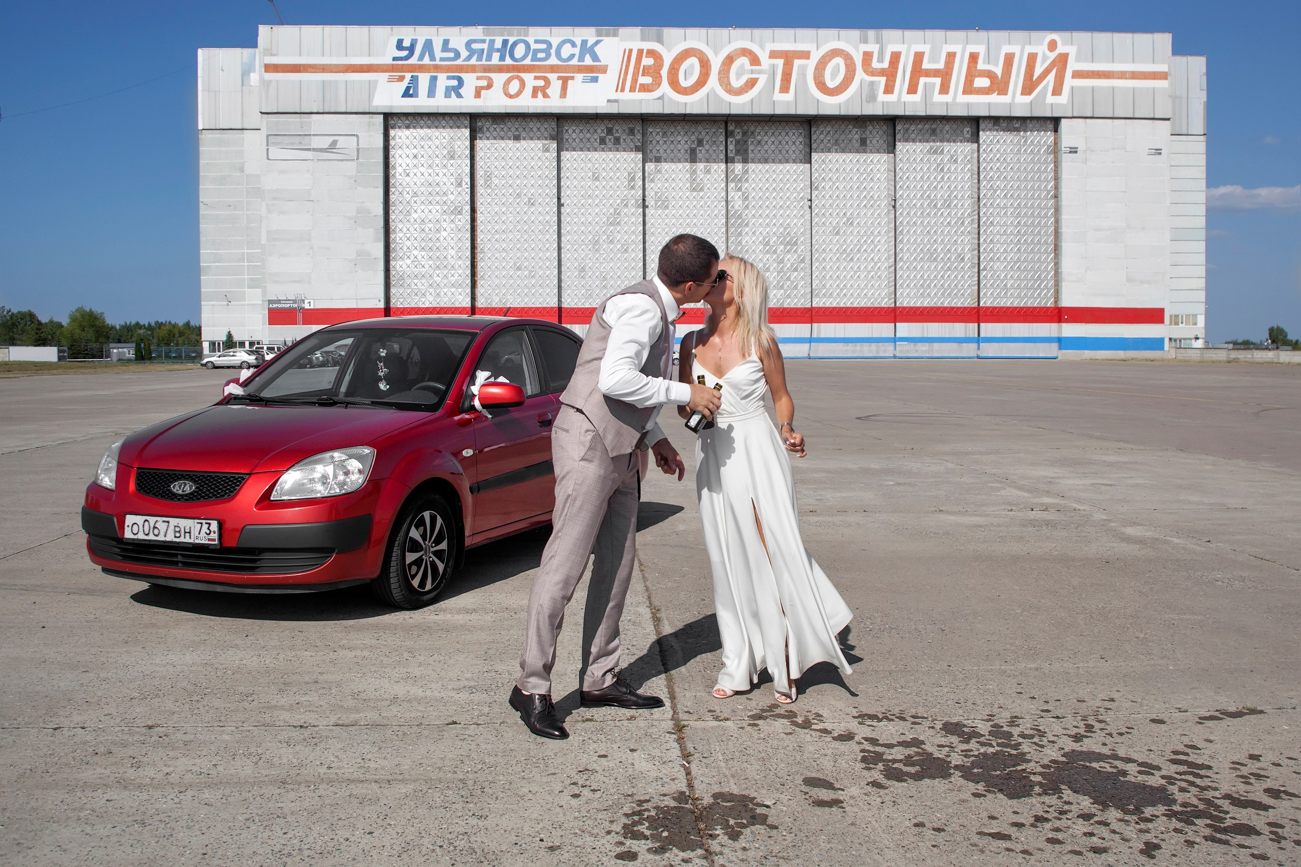 Жених с невестой целуются на фоне красной машины в аэропорту
