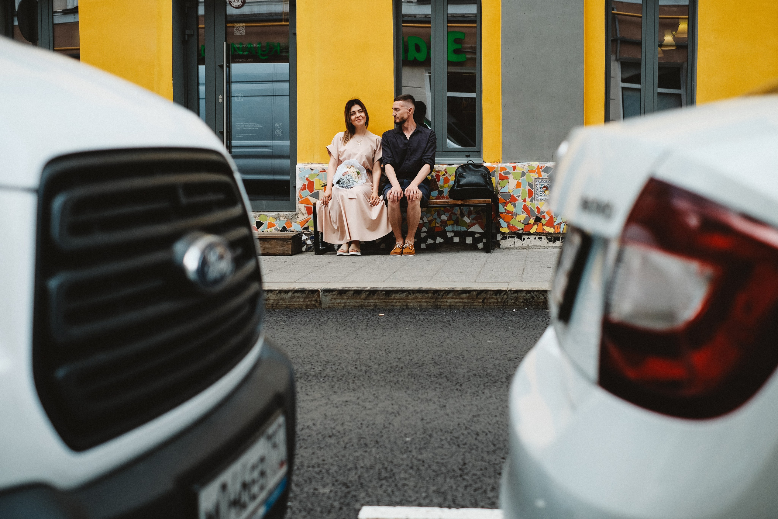 Пара сидит на скамейке, съёмка между машинами.