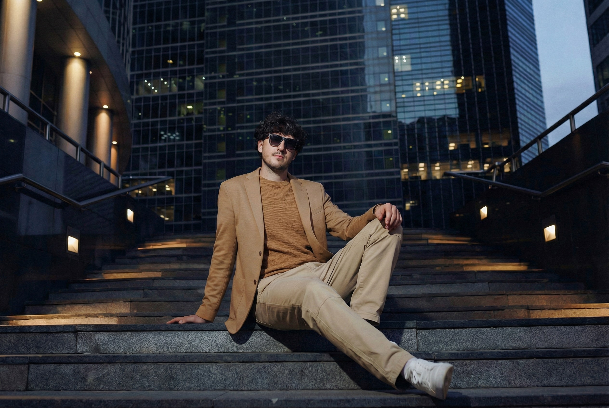 Стильный портрет в тёмных очках: мужчина сидит на лестнице, наблюдая за закатом над небоскрёбами Москва-Сити. Атмосферный кадр.
