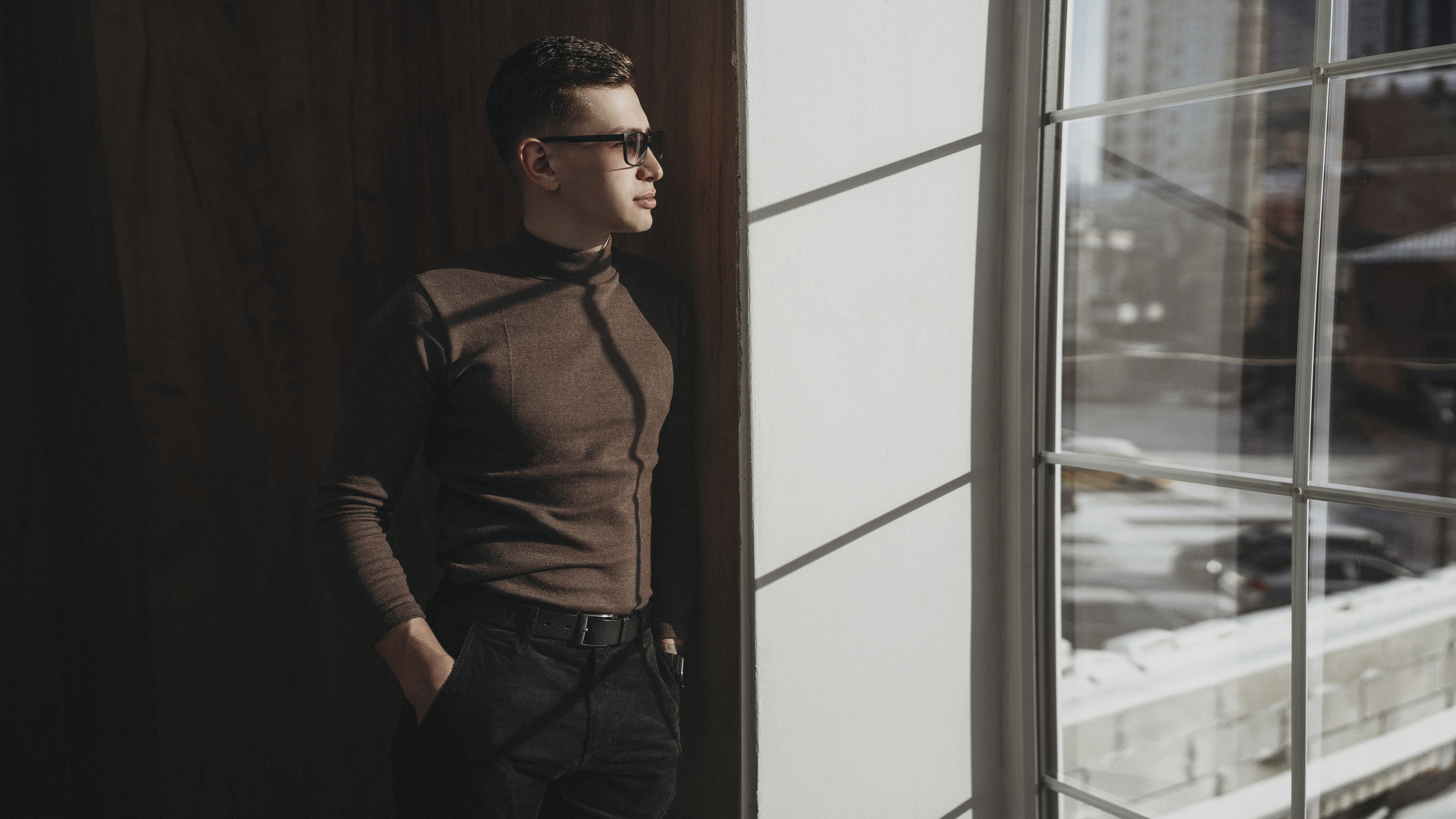 Брутальный парень в очках стоит у окна и смотрит в окно.