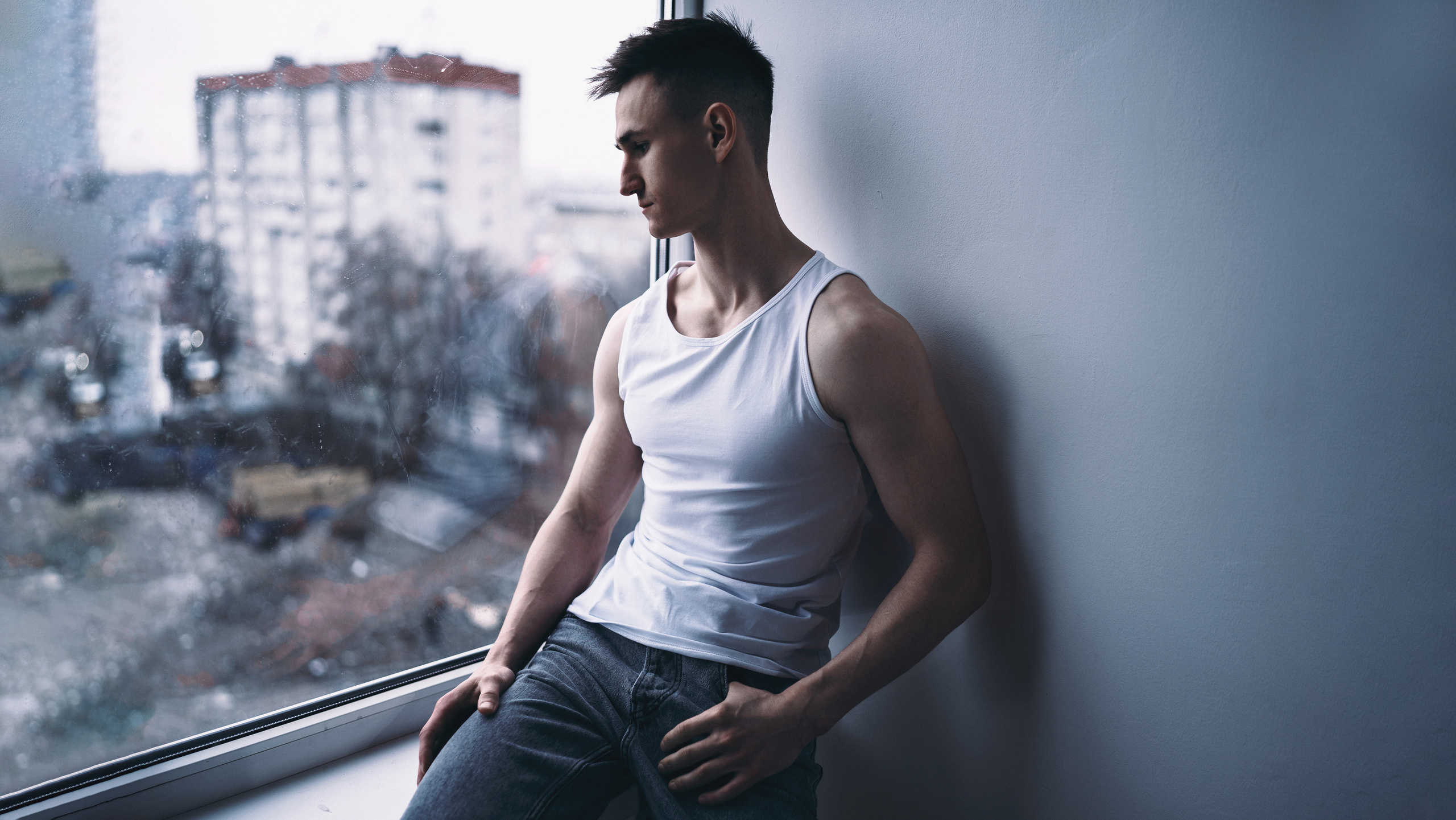 Парень в белой майке сидит на подоконнике и смотрит вдаль.