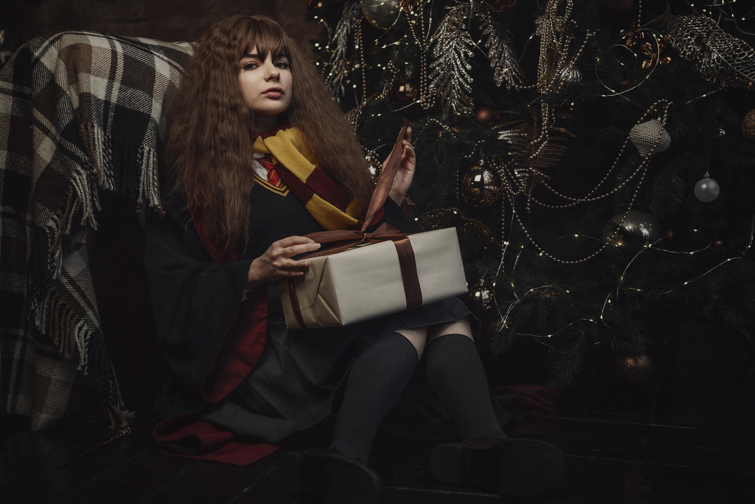 Девушка в образе Гермионы с подарком сидит у елки.