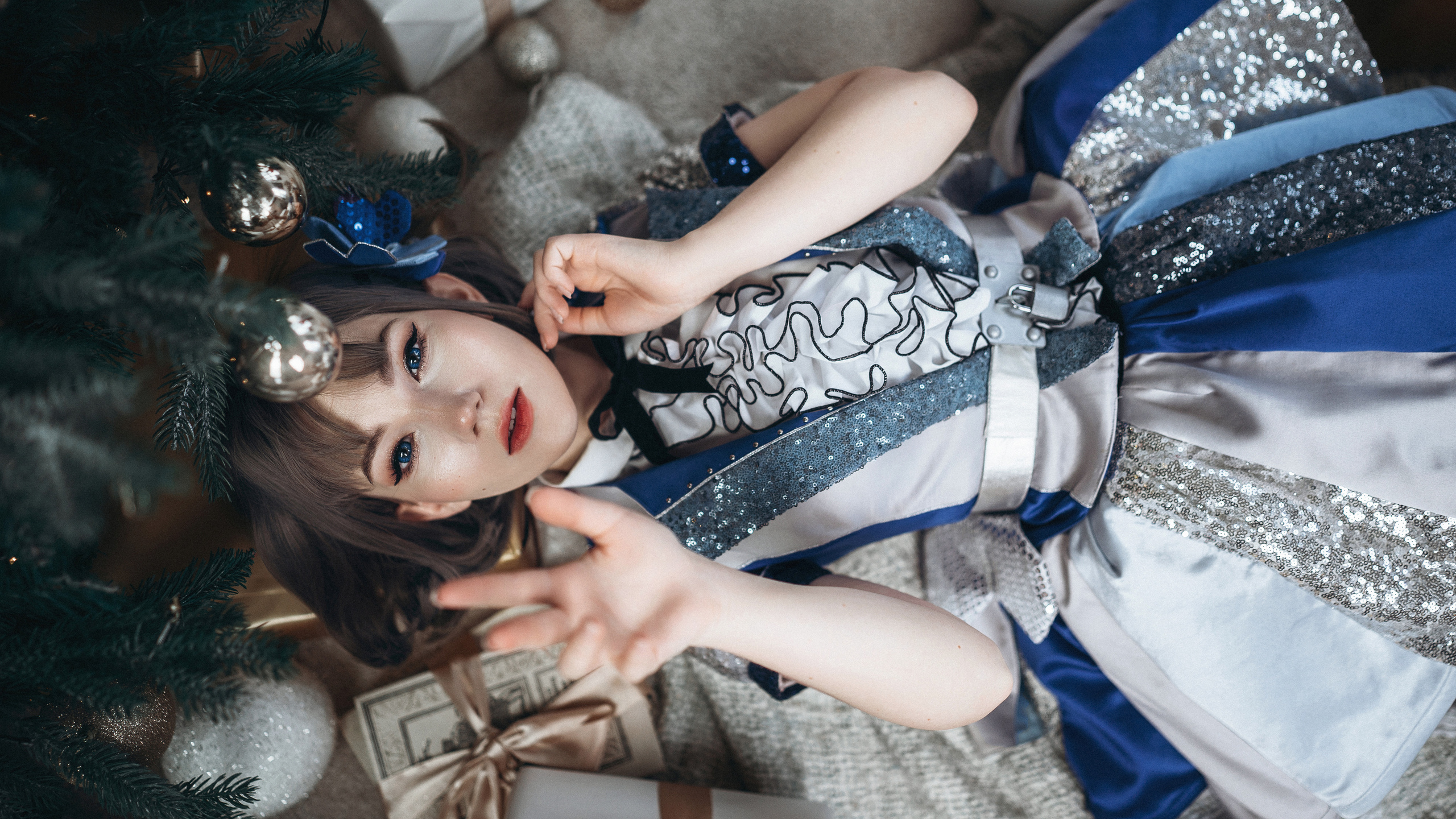 Девушка в синем платье лежит на полу у елки и тянет руку вверх.