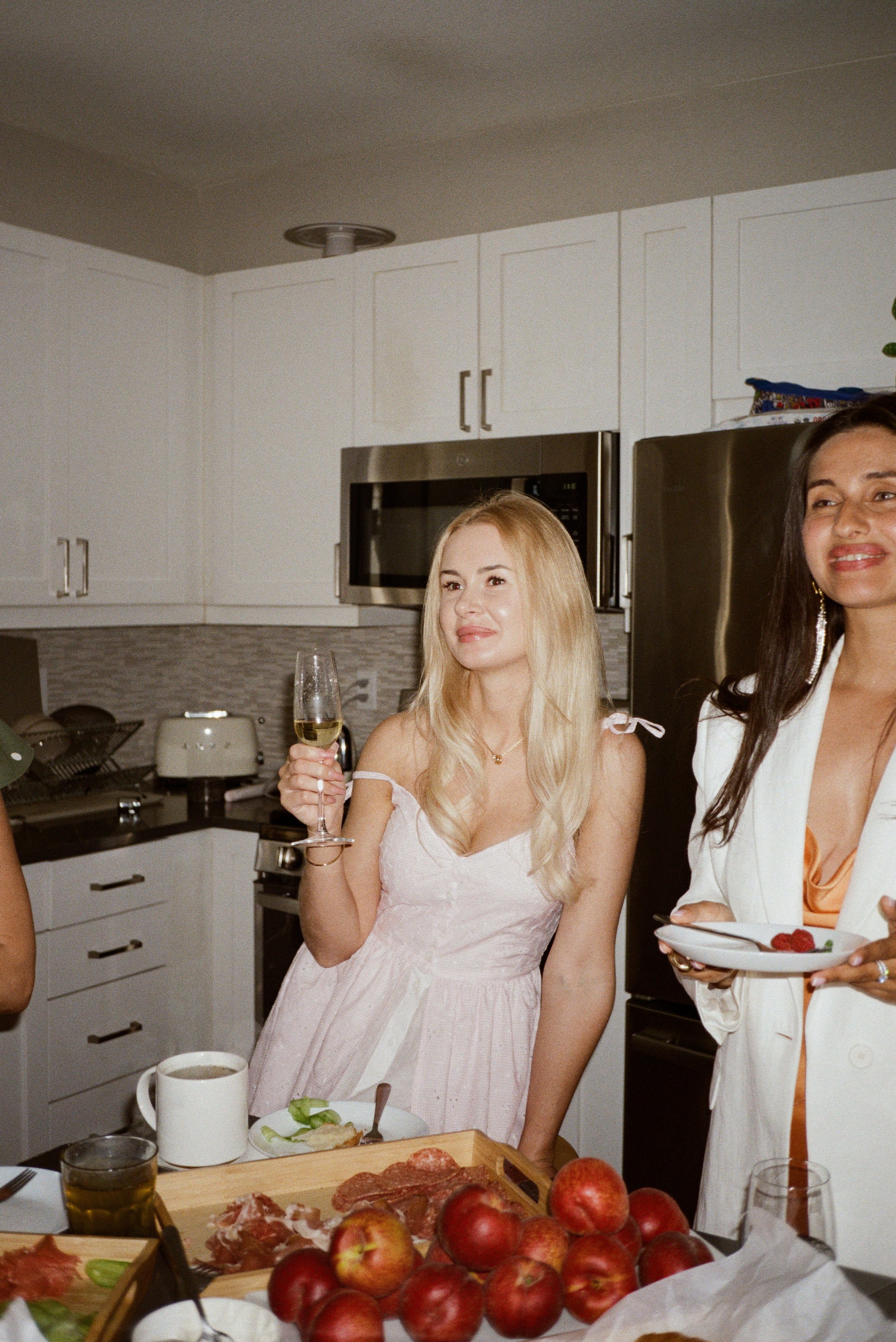 A candid film photo taken in the kitchen of a woman with blond hair in pink dress with champagne glass 