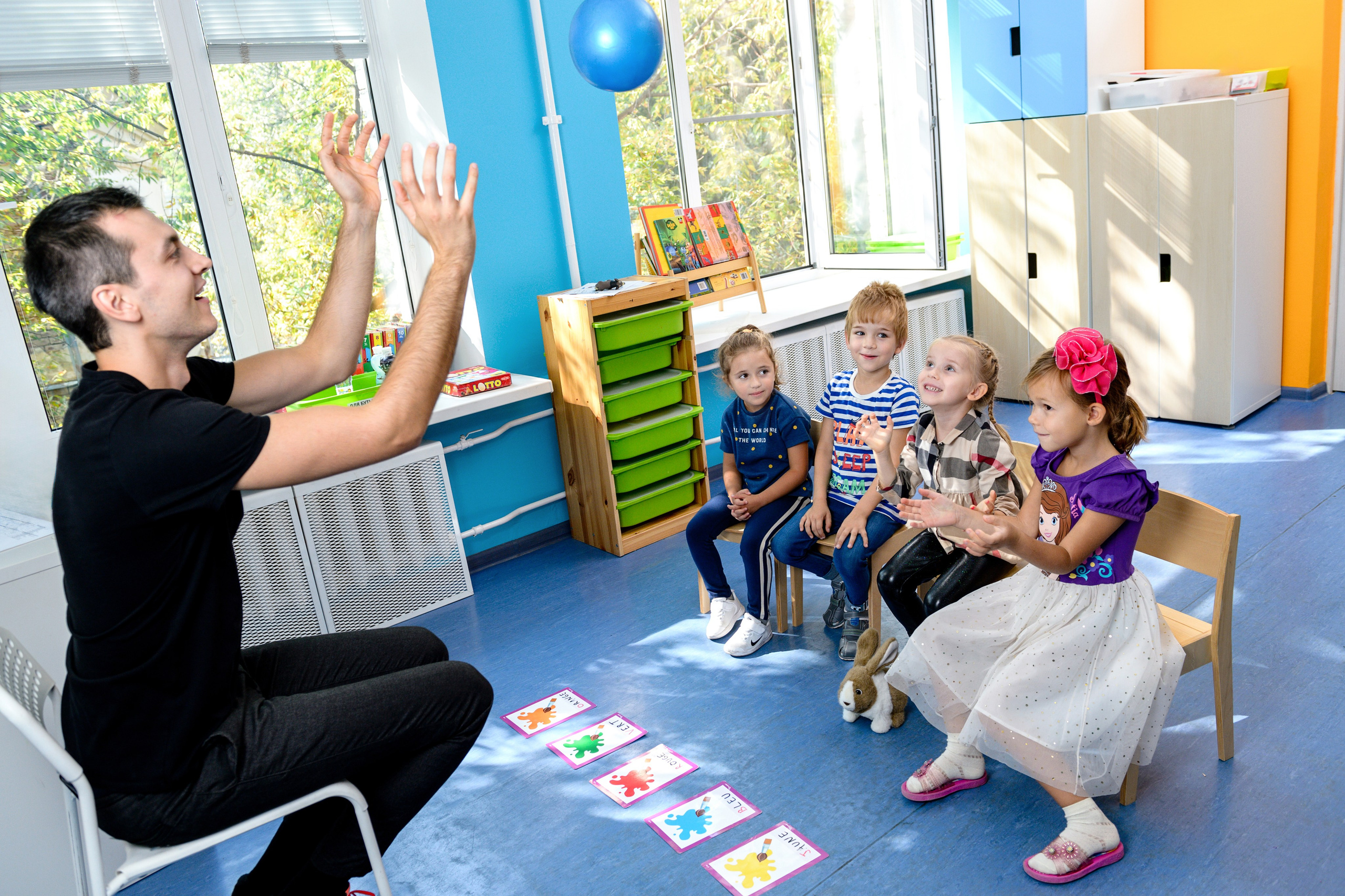 Воспитатель на занятии в детском саду показывает движения, дети сидят в ряд и повторяют в светлом игровом классе.