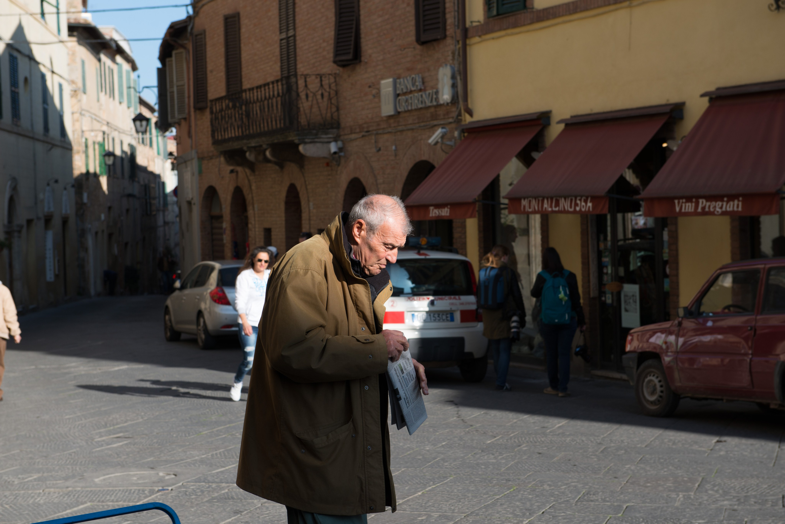 Мужчина в коричневом пальто в фокусе читает газету на солнечной улице в Италии с мягкими тенями.