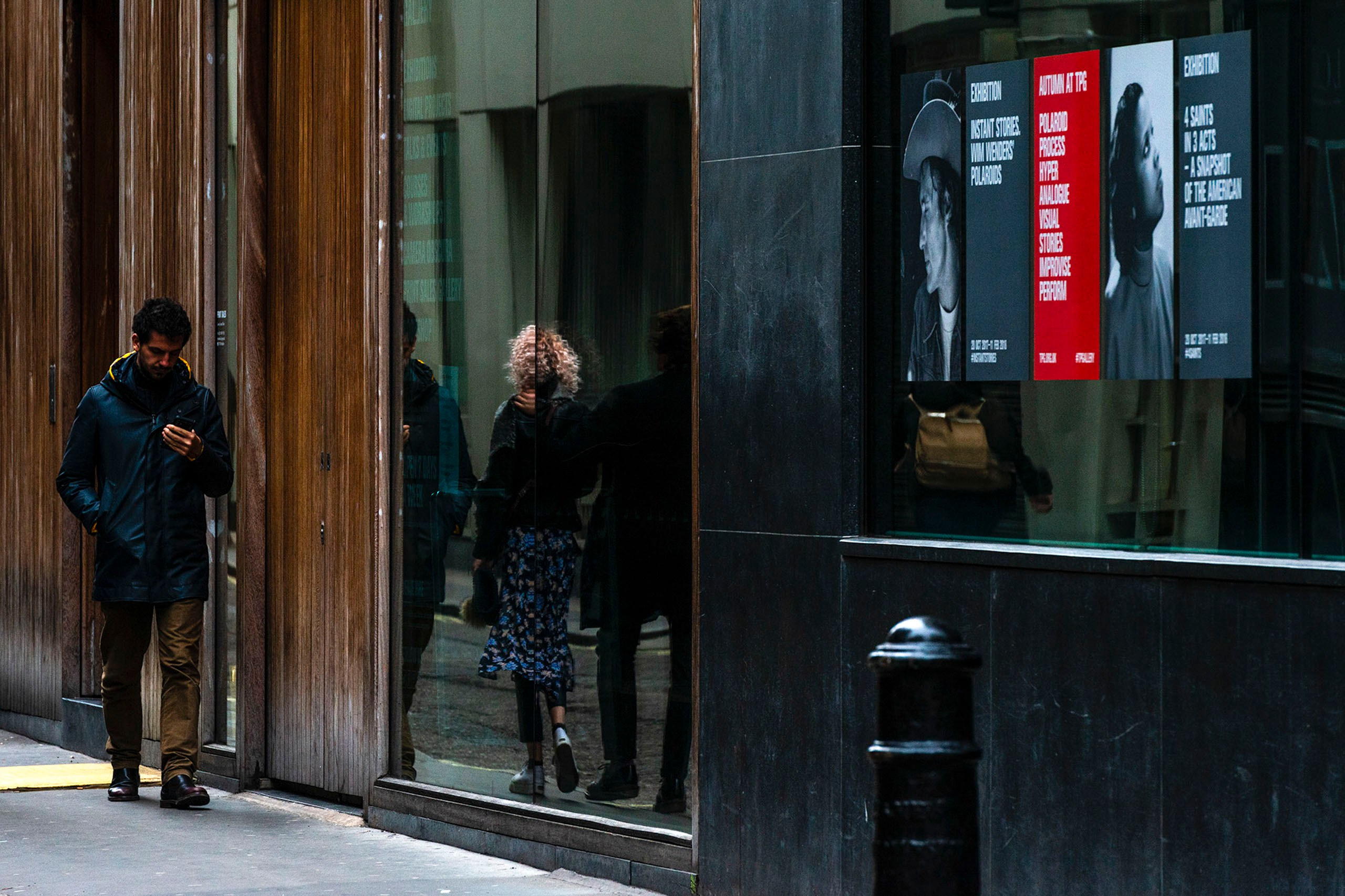 Стрит-фотография мужчины в синей куртке, смотрящего в телефон на фоне витрины с отражением женщины