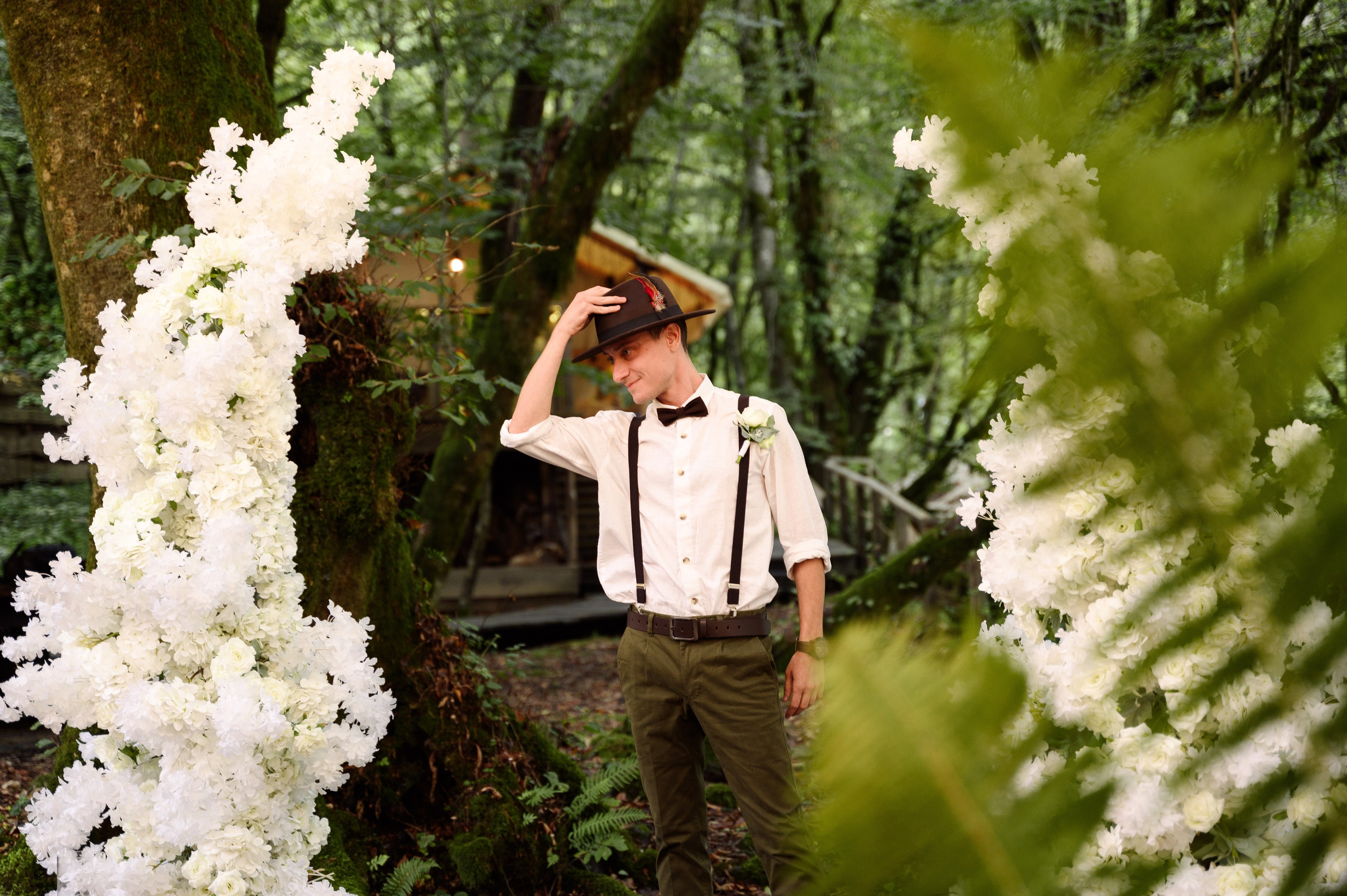 Жених стоит у свадебной арки и только только хочет снять шляпу на фотограф Сочи успел за впечатлить момент когда он ещё не успел снять шляпу на фоне лес и на переднем плане ветка расфокусе