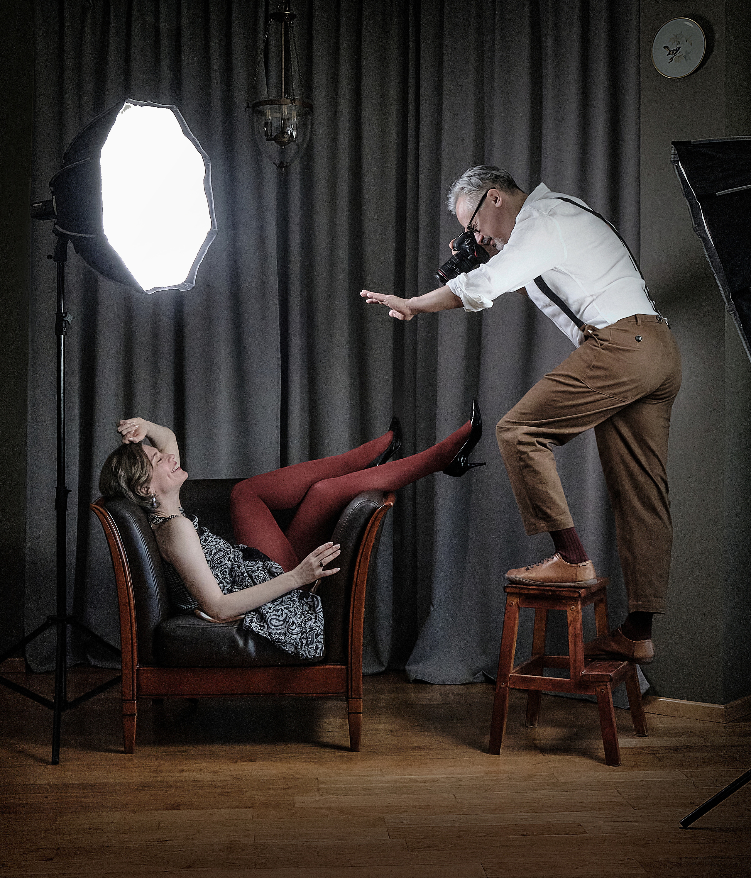 Мужчина в фотостудии фотографирует женщину в платье на кресле и жестом просит ее не двигаться 