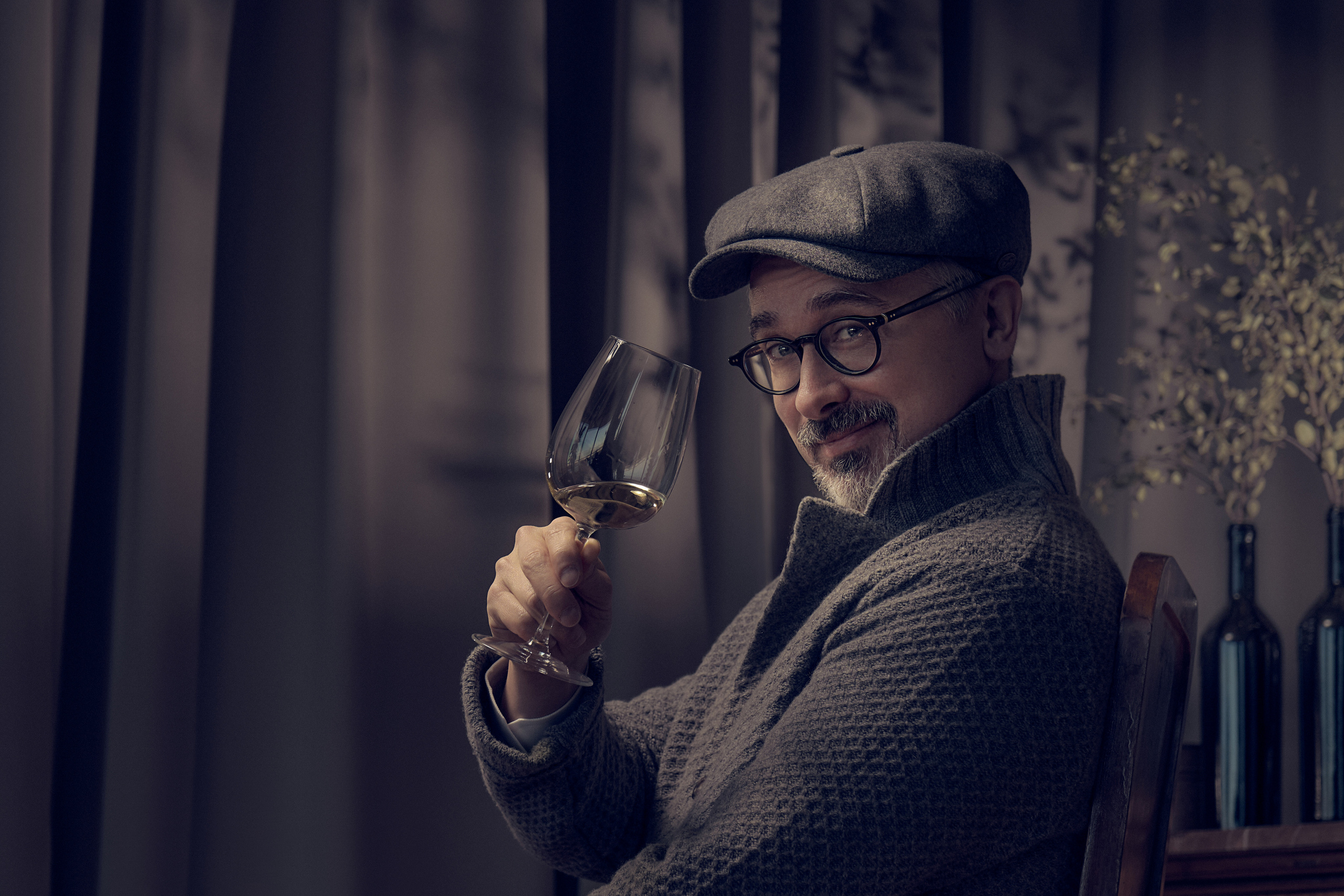 Мжчина дегустатор с седой бородой держит в руке бокал вина жестом предлагая выпить с ним