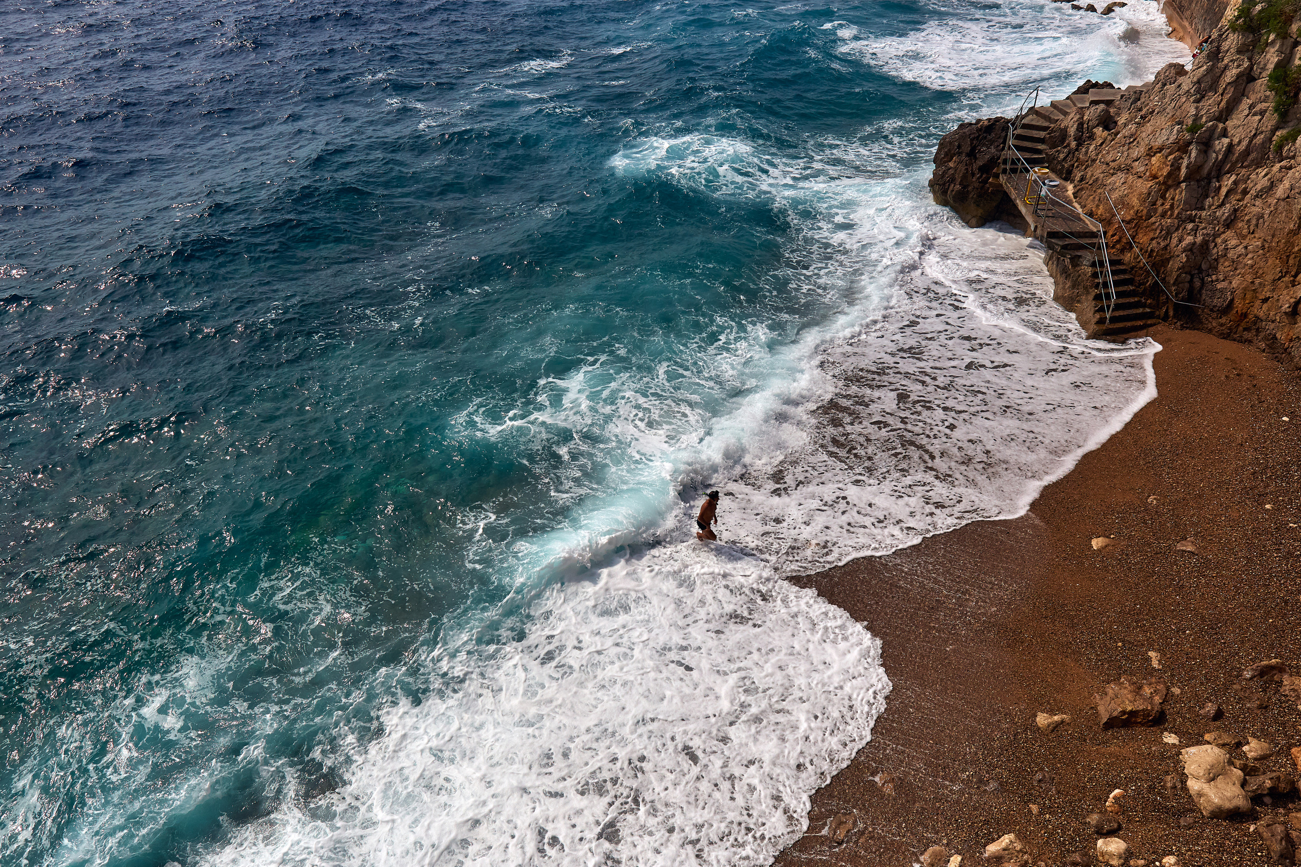 Лазурное побережье с небольшим пляжем и пеной волн из которых на берег выходит отдыхающий турист