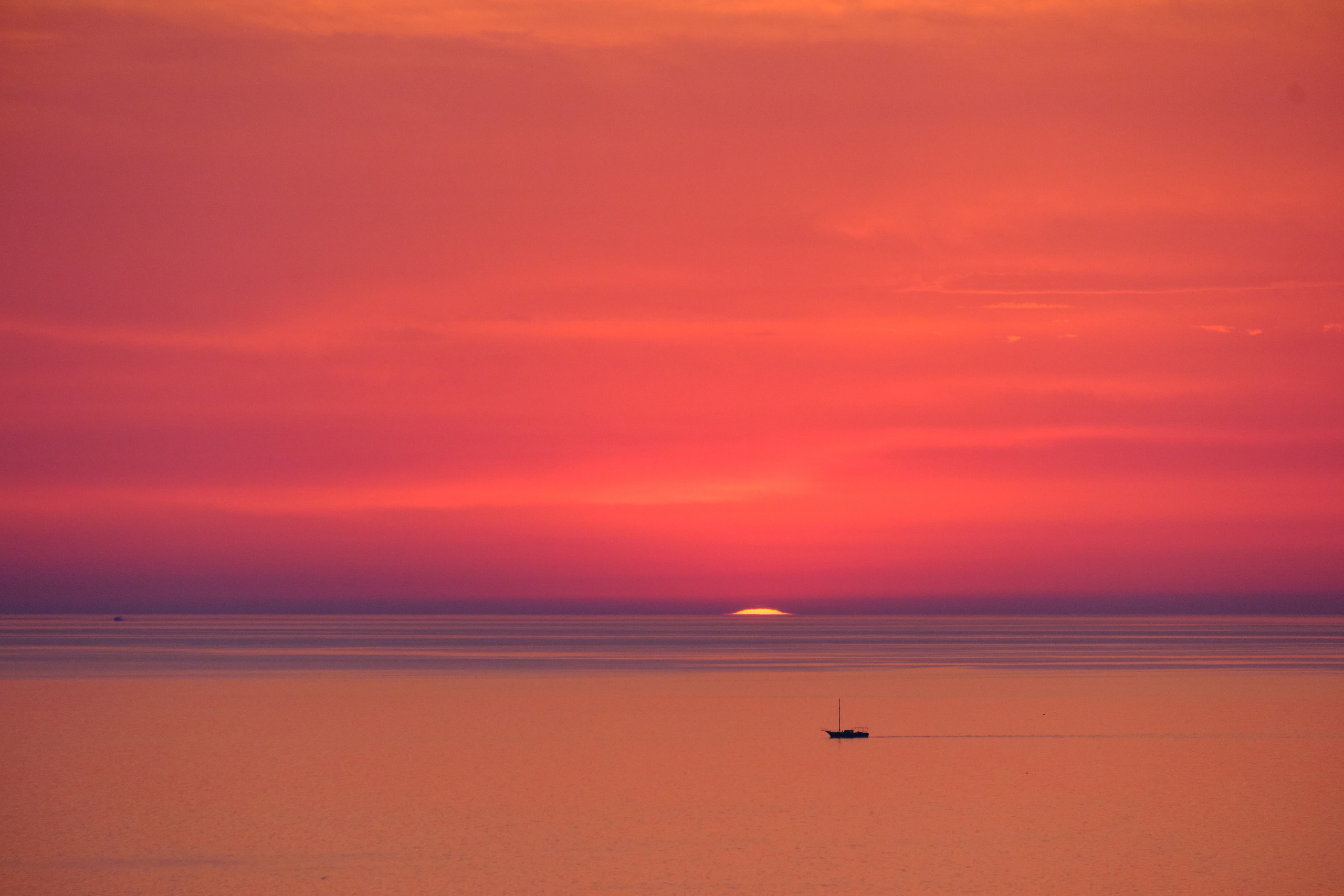 Красный закат, где солнце почти скрылось за морским горизонтом, а по водной глади плывет небольшой корабль