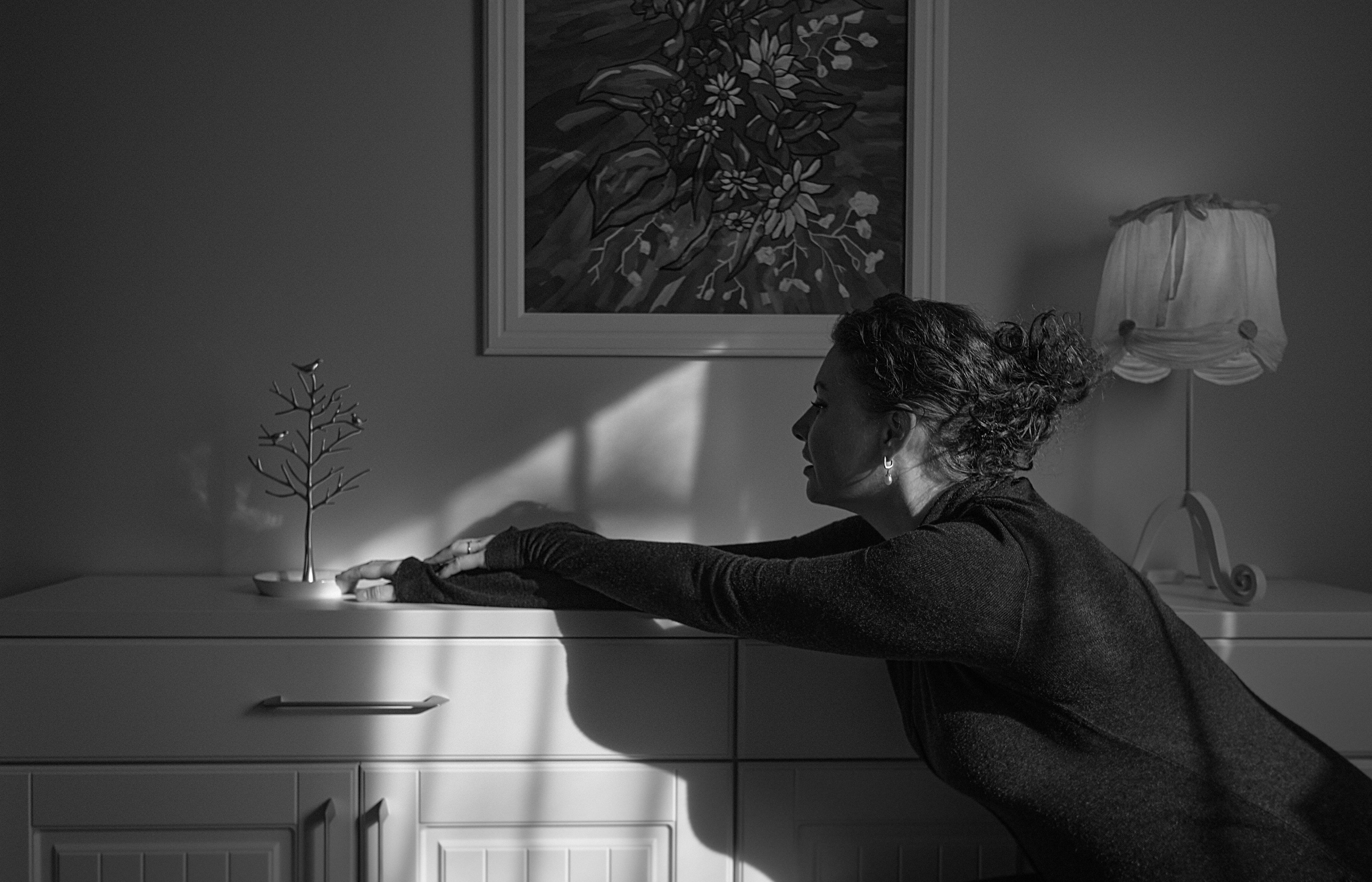 Женщина в черной кофте освещена светом из окна сидит у белого комода, на котором стоит настольная лампа и видна картина на стене