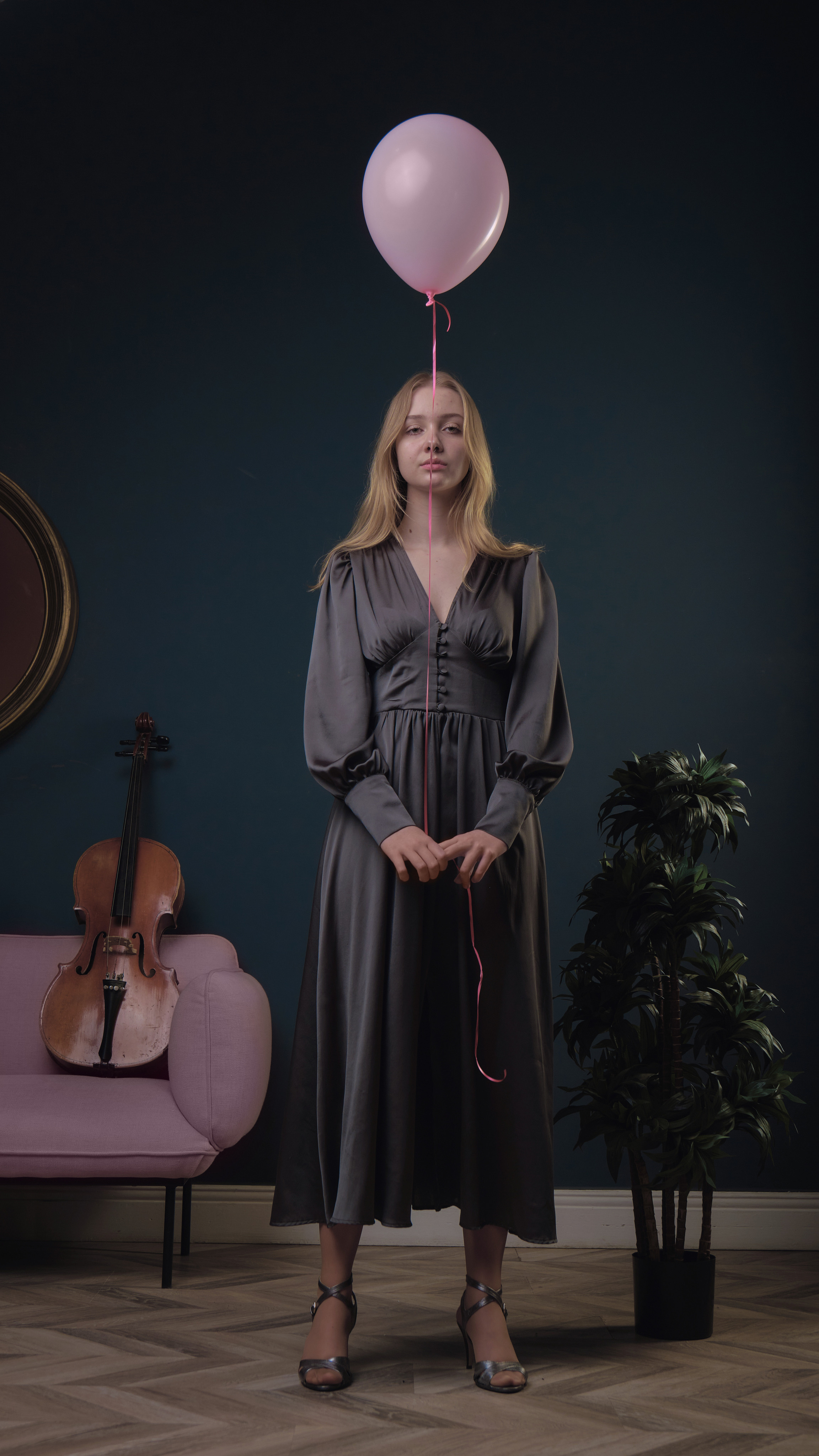 Белокурая девушка на темном фоне в темном платье держит розовый шарик над головой 