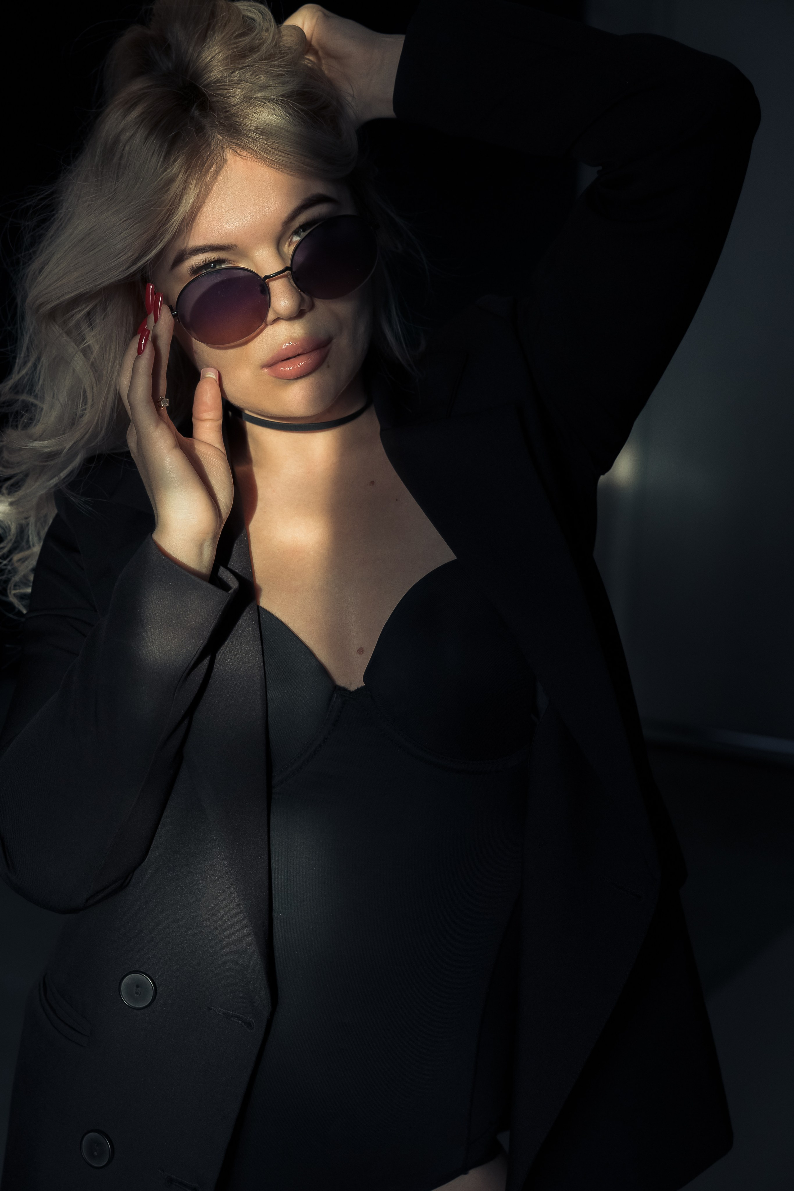 Девушка в тёмных очках и пальто, позирует на тёмном фоне, загадочный образ.