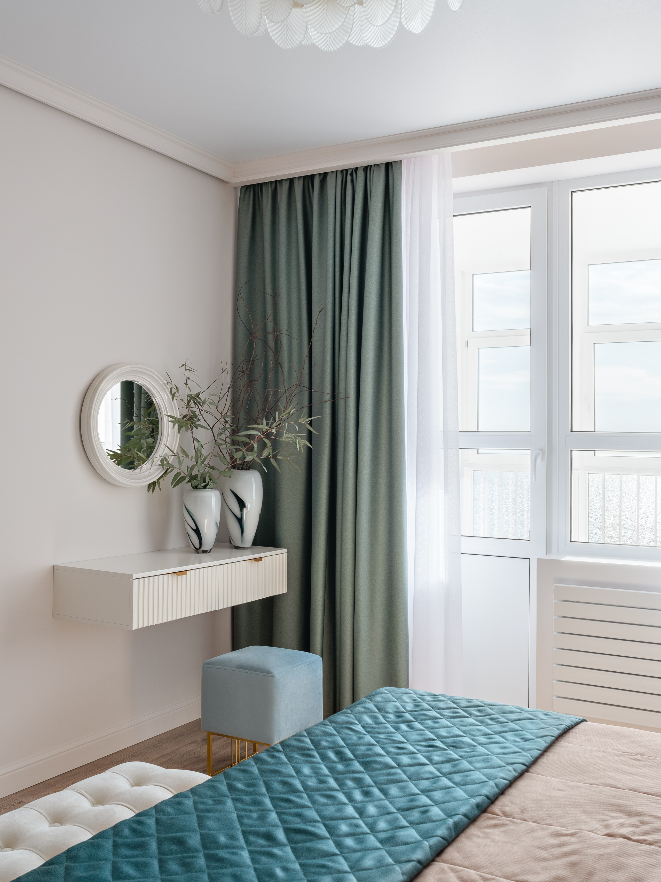 Светлая спальня с большим окном, зелёными портьерами, белыми тюлями и минималистичной подвесной консолью с зеркалом, дополненной декоративными вазами. Интерьер создан дизайнером Натальей Бунеевой, стилистика от Натальи Якушевой, фотосъёмка Сергея Денисенко.