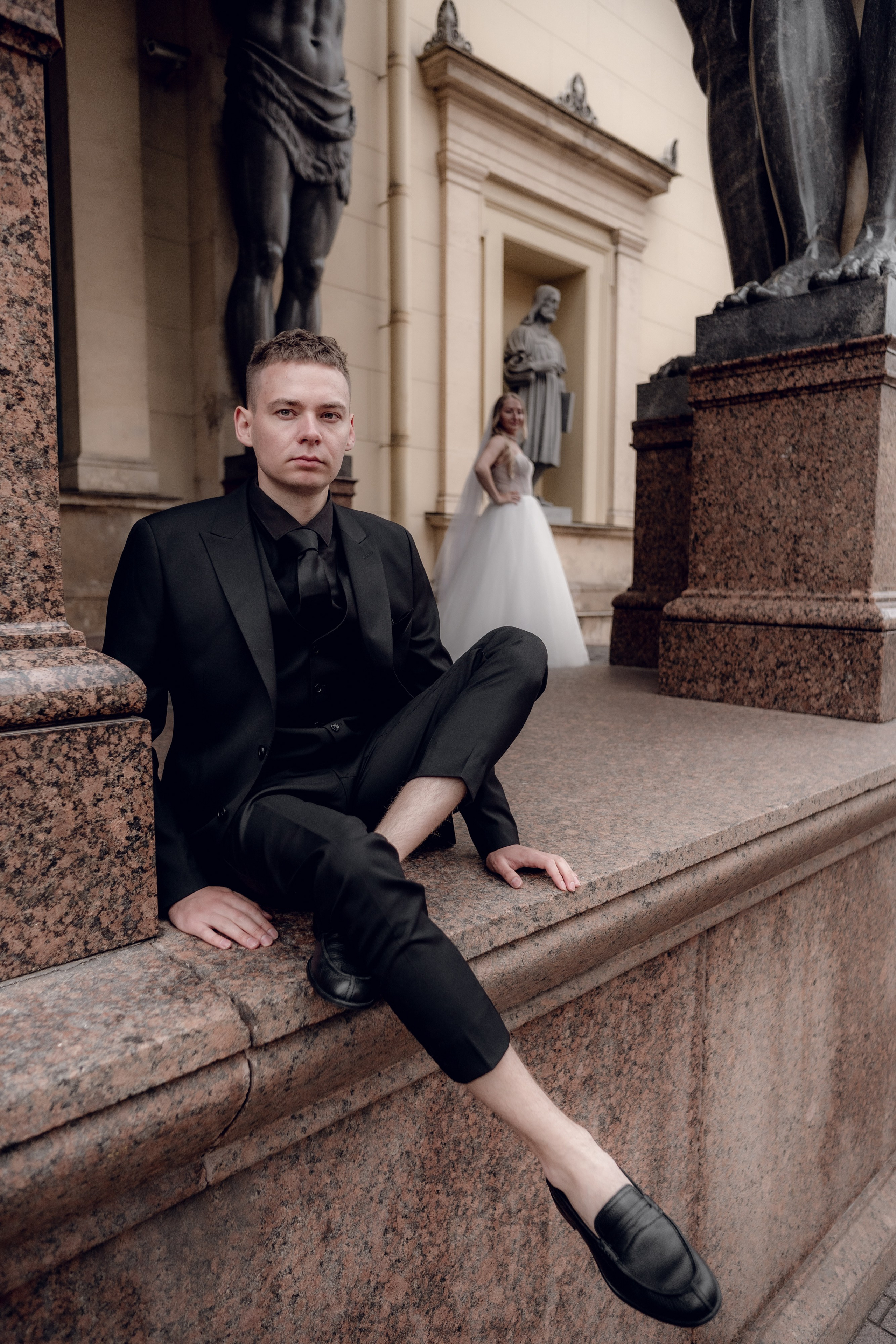 передний план - жених в черном костюме в укороченных брюках, задний план - стоит невеста около атлантов