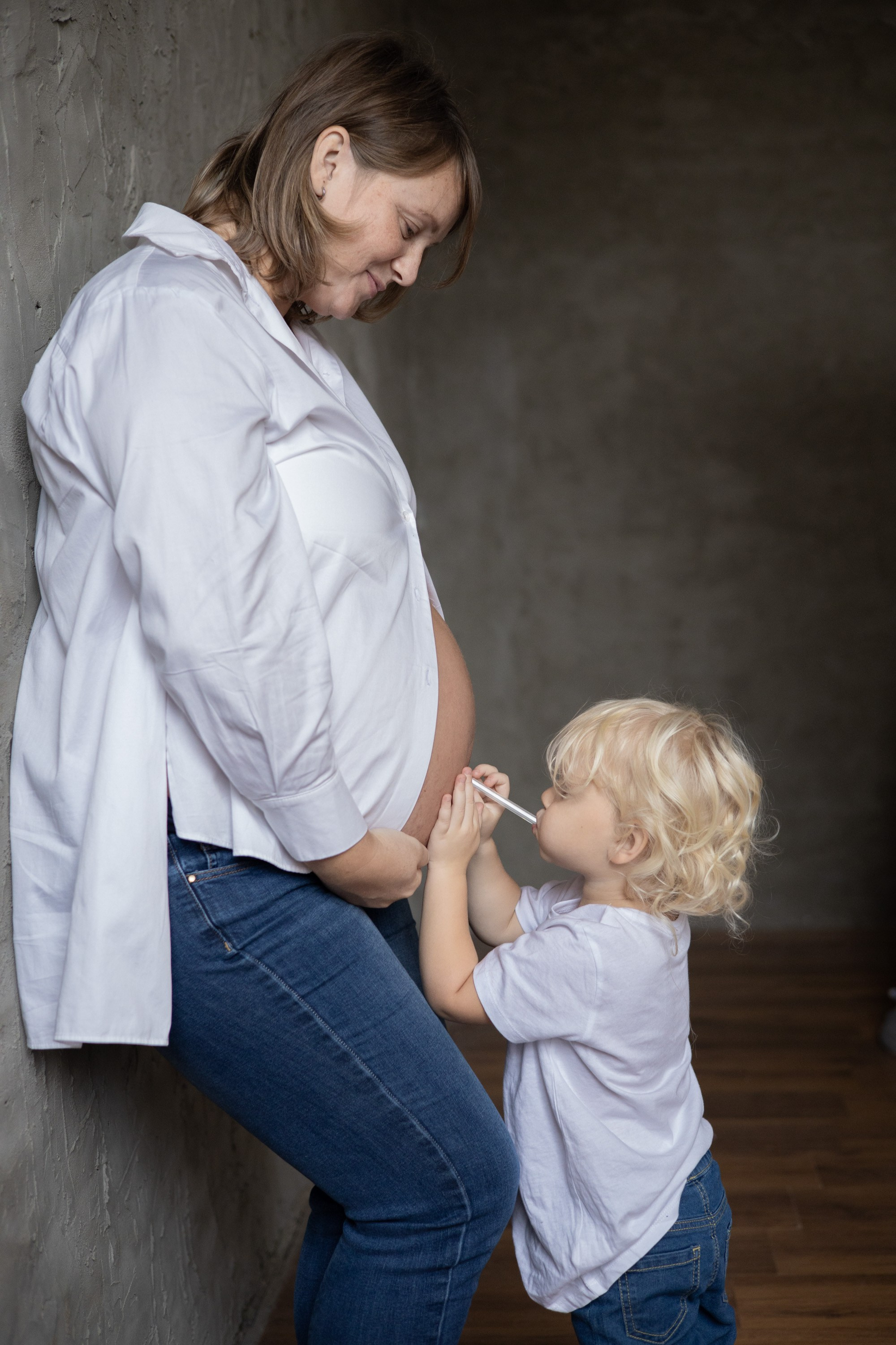 Маленький мальчик "надувает" мамин живот через соломинку. Беременная мама стоит у стены. Фотосессия для беременных  Астрахань.