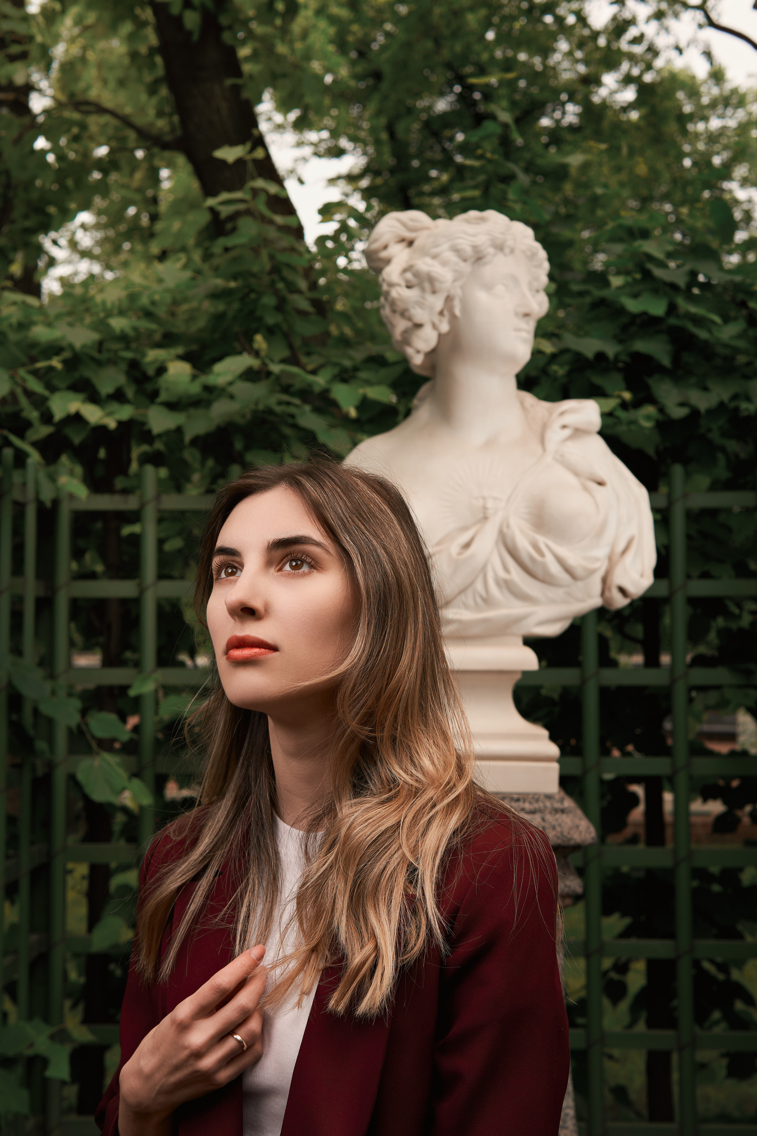 прогулочная фотосессия девушки в бордовом костюме в летнем саду со вспышкой летом в зелени в Санкт-Петербурге со статуей в стиле кино, стробизм днем в пасмурную погоду