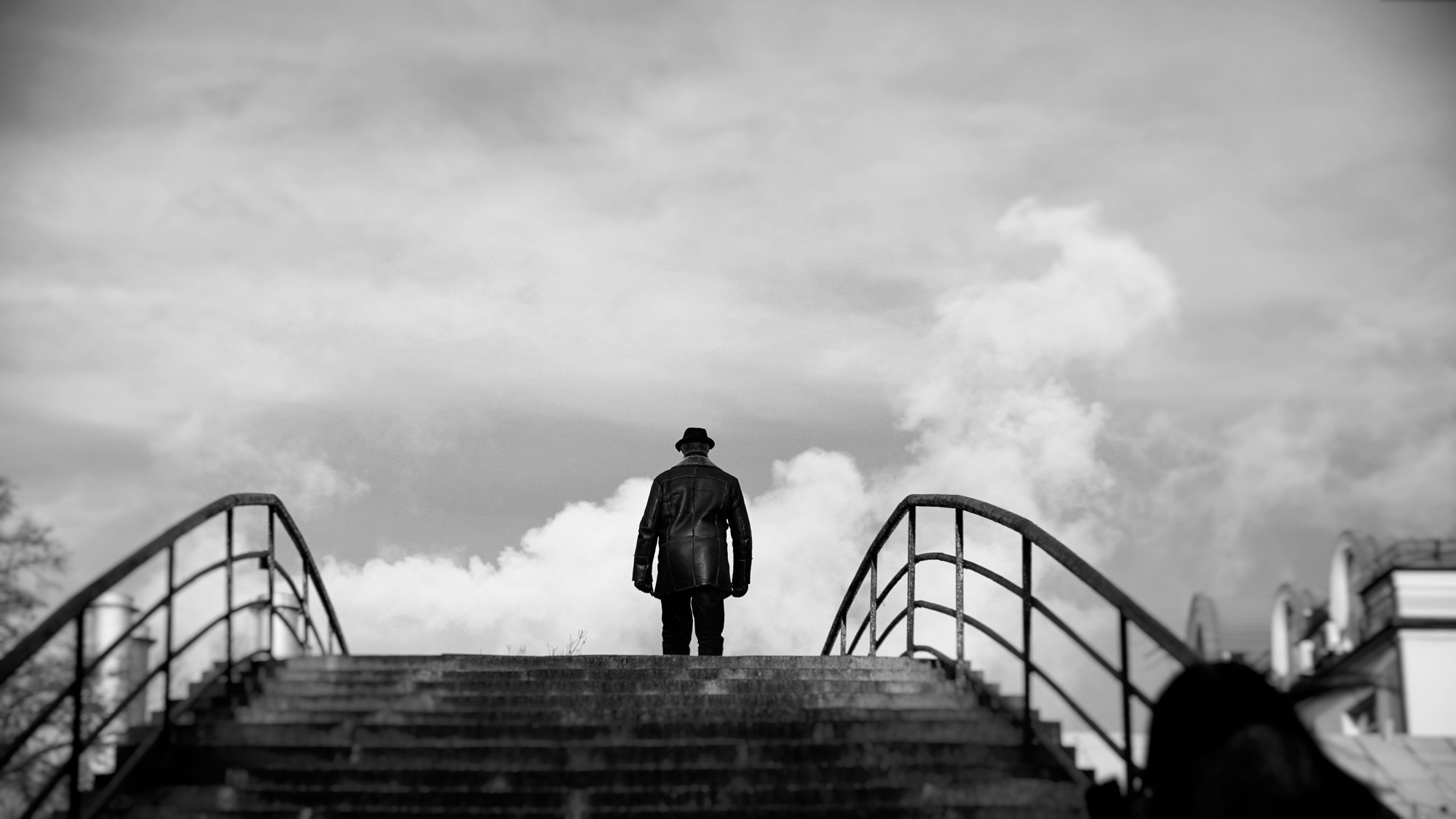 Черно-белое фото человека в кожаной куртке и шляпе стоящего на мосту спиной к камере, драматичное небо с облаками, философский образ