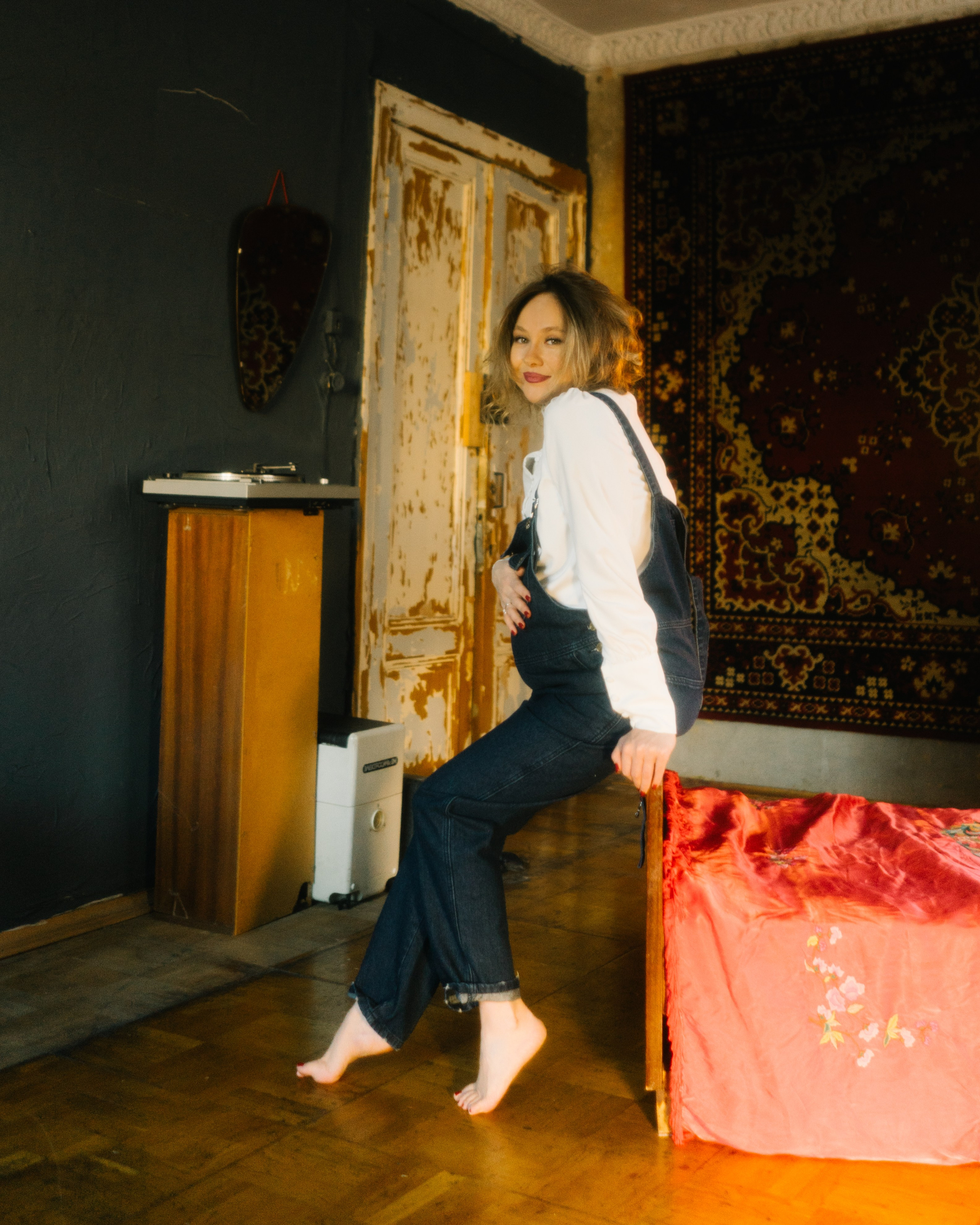 Беременная в белой блузе и темном комбинезоне у кровати — стильная фотосъемка беременности в винтажном интерьере.