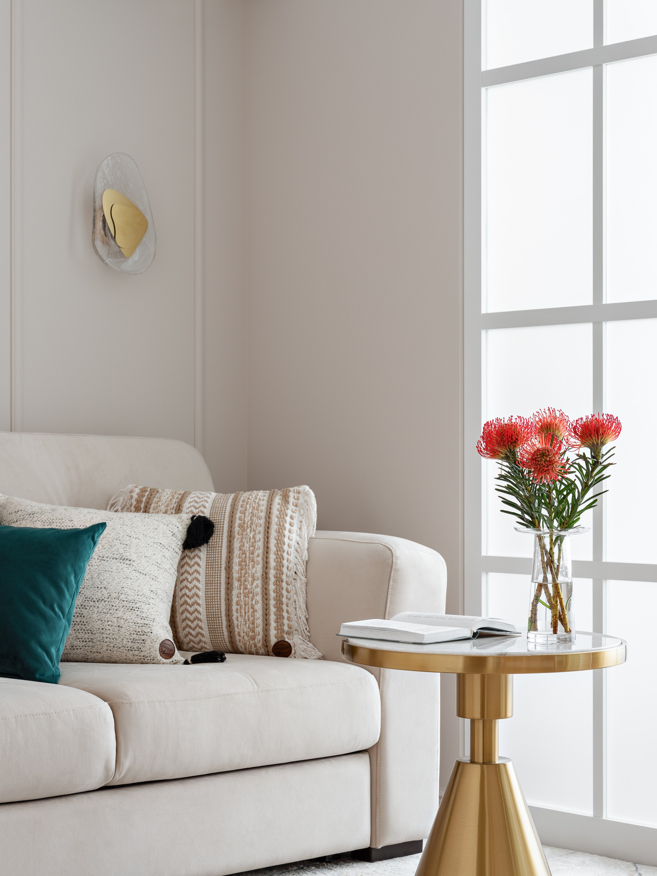 Уютный фрагмент гостиной с мягким диваном в светлой обивке, декоративными подушками и стильным круглым столиком с золотым основанием. На столе — ваза с цветами и книга. Интерьер: дизайнер Наталья Бунеева, стилист Наталья Якушева, фотограф Сергей Денисенко.