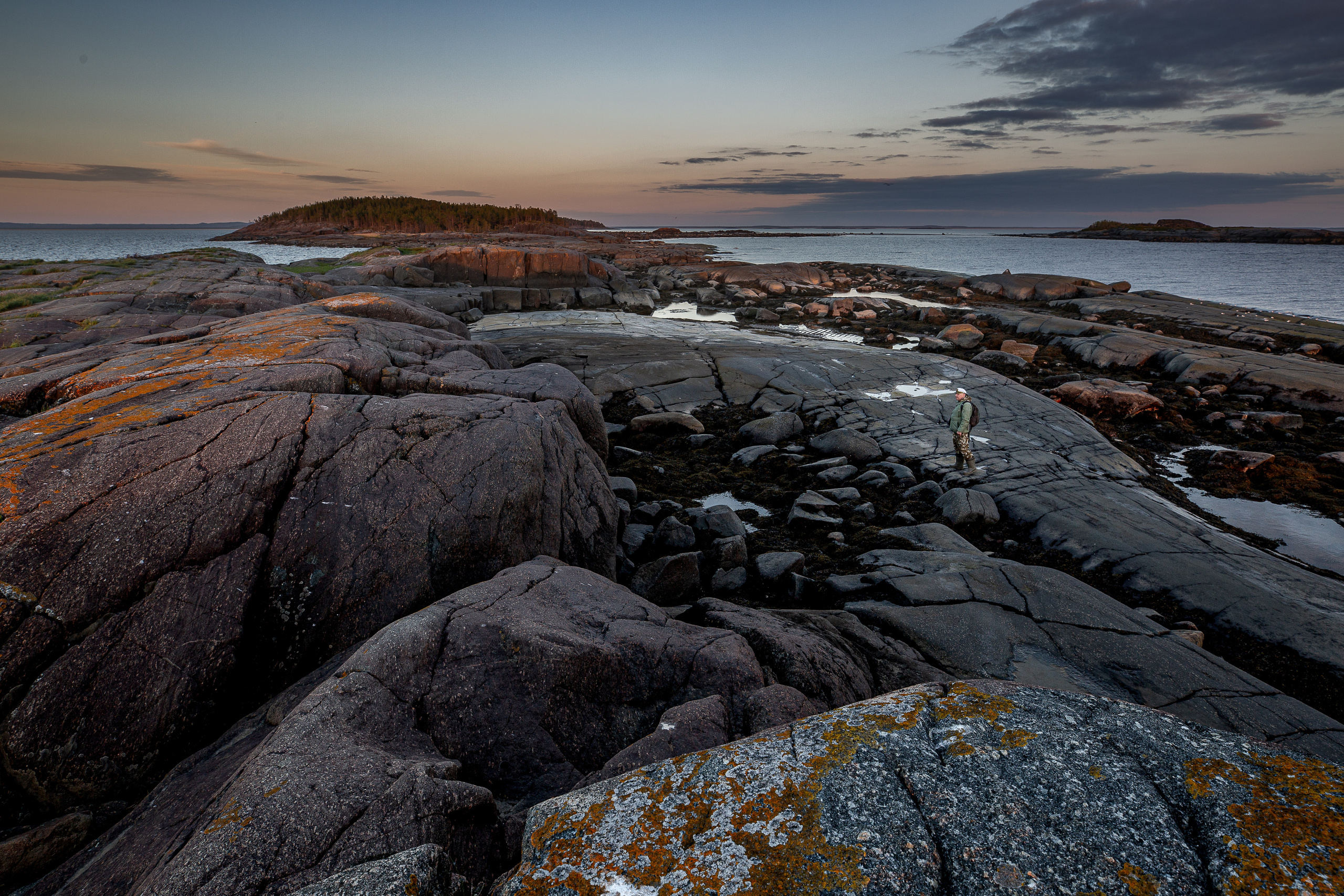 Одинокий человек в зелёной куртке и кепке стоит камнях Кий острова на фоне  вечернего неба. 
