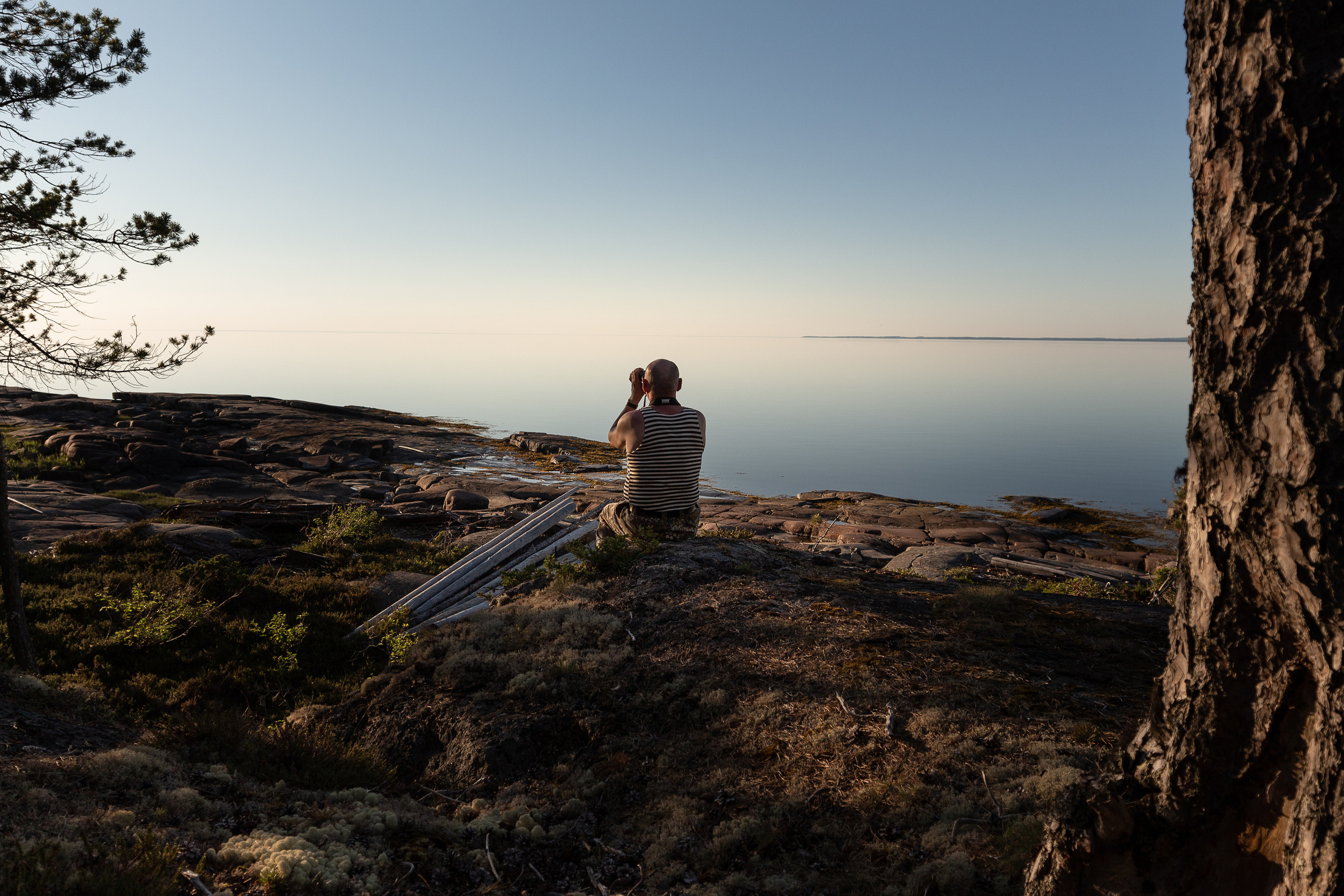 Мужчина с биноклем сидит в тельняшке смотрит вдаль. 
