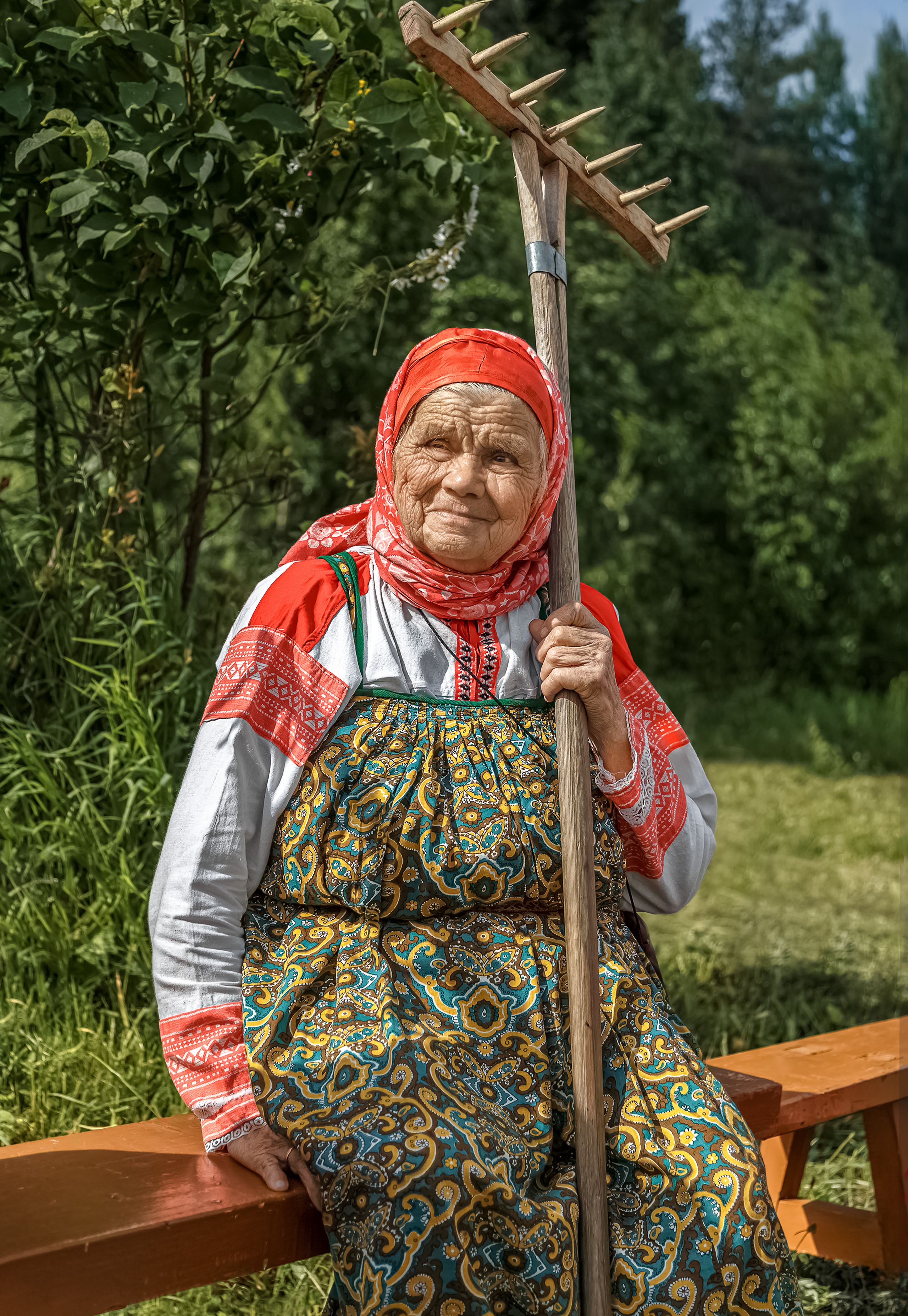 Бабушка в красном платке и русском  красном сарафане с граблями.