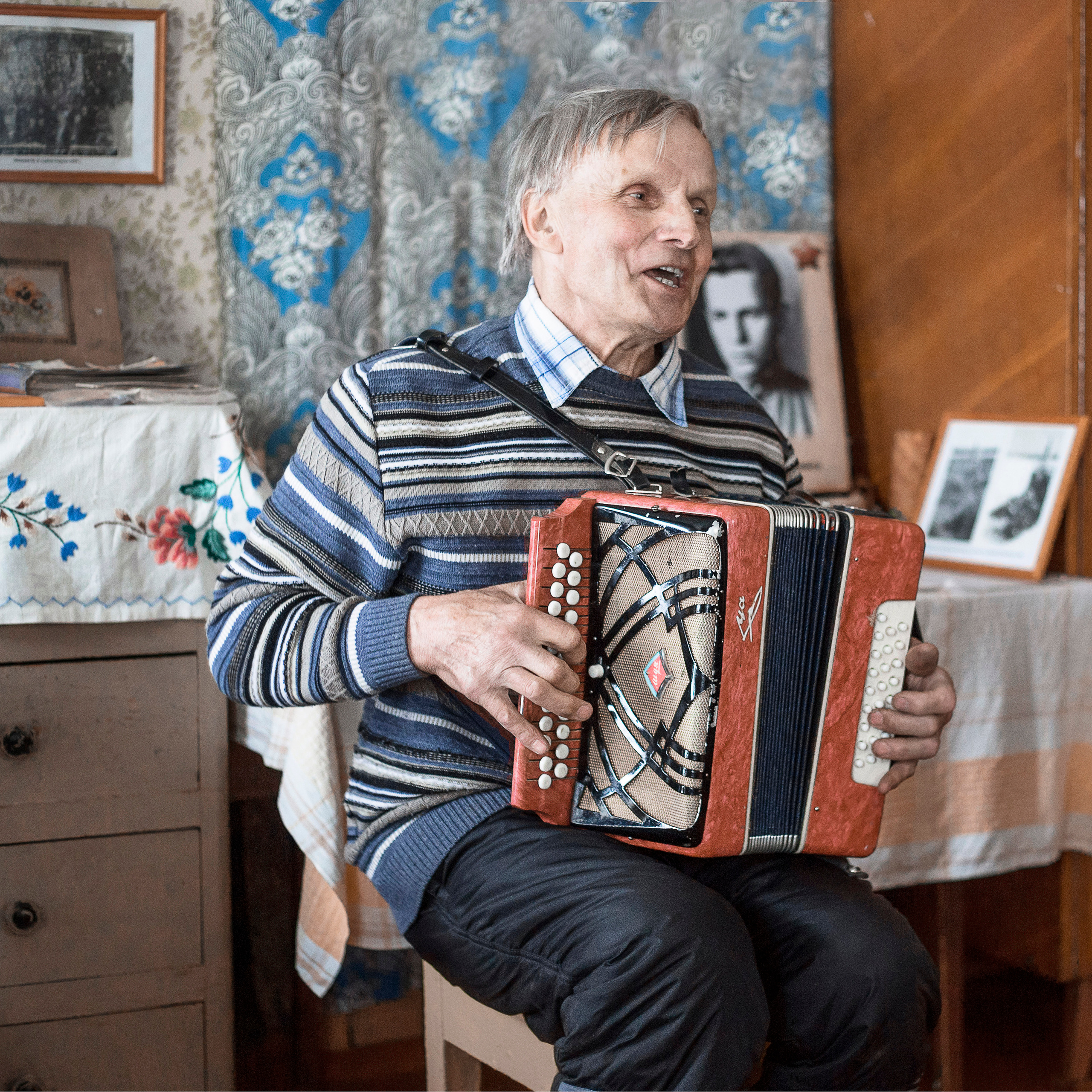 Мужчина играет на гармони и поет в деревенском доме.
