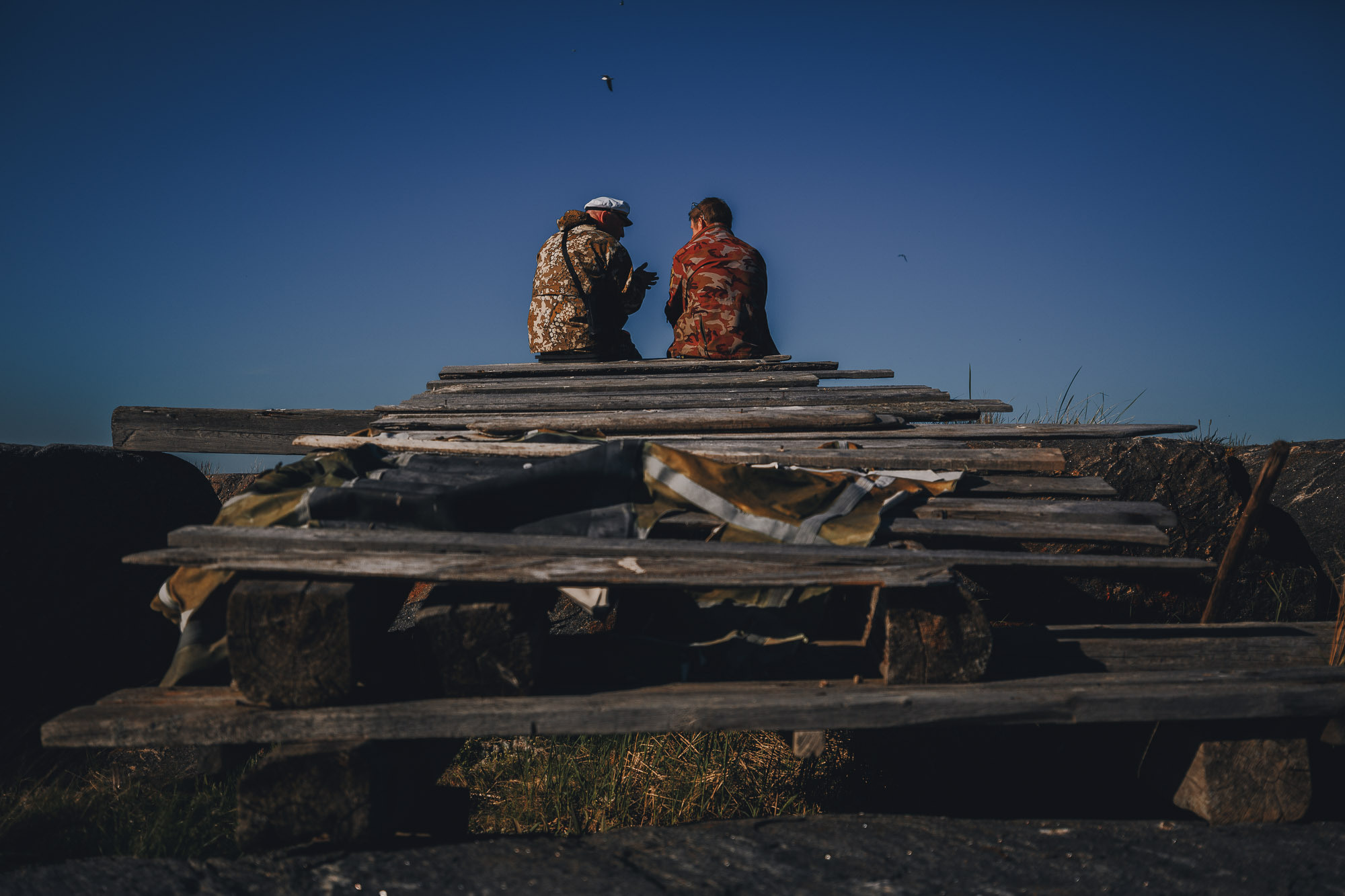 Двое мужчин в рыбацкой одежде сидят на деревянных мостках и беседуют на фоне синего неба на острове. 