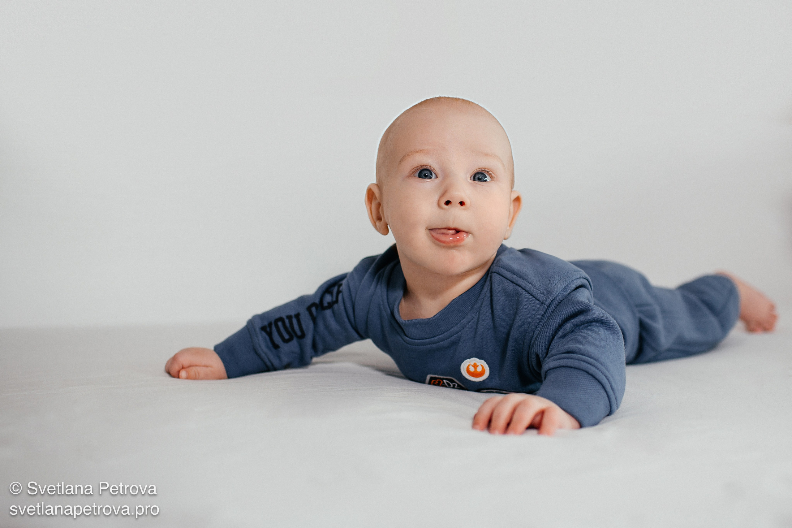 Младенец на животе, игривый взгляд в камеру. Любопытный малыш в синем костюме. Ребёнок на светлом фоне, поза «ползунка». Малыш с высунутым язычком, студийный кадр.Студия, младенец, весёлый взгляд. Ребёнок в расслабленной позе на фото. Улыбающийся малыш на светлом фоне. Фотосессия младенца: поза на животе. Студия, малыш в синем, игривое настроение. Младенец с озорным взглядом в кадре.