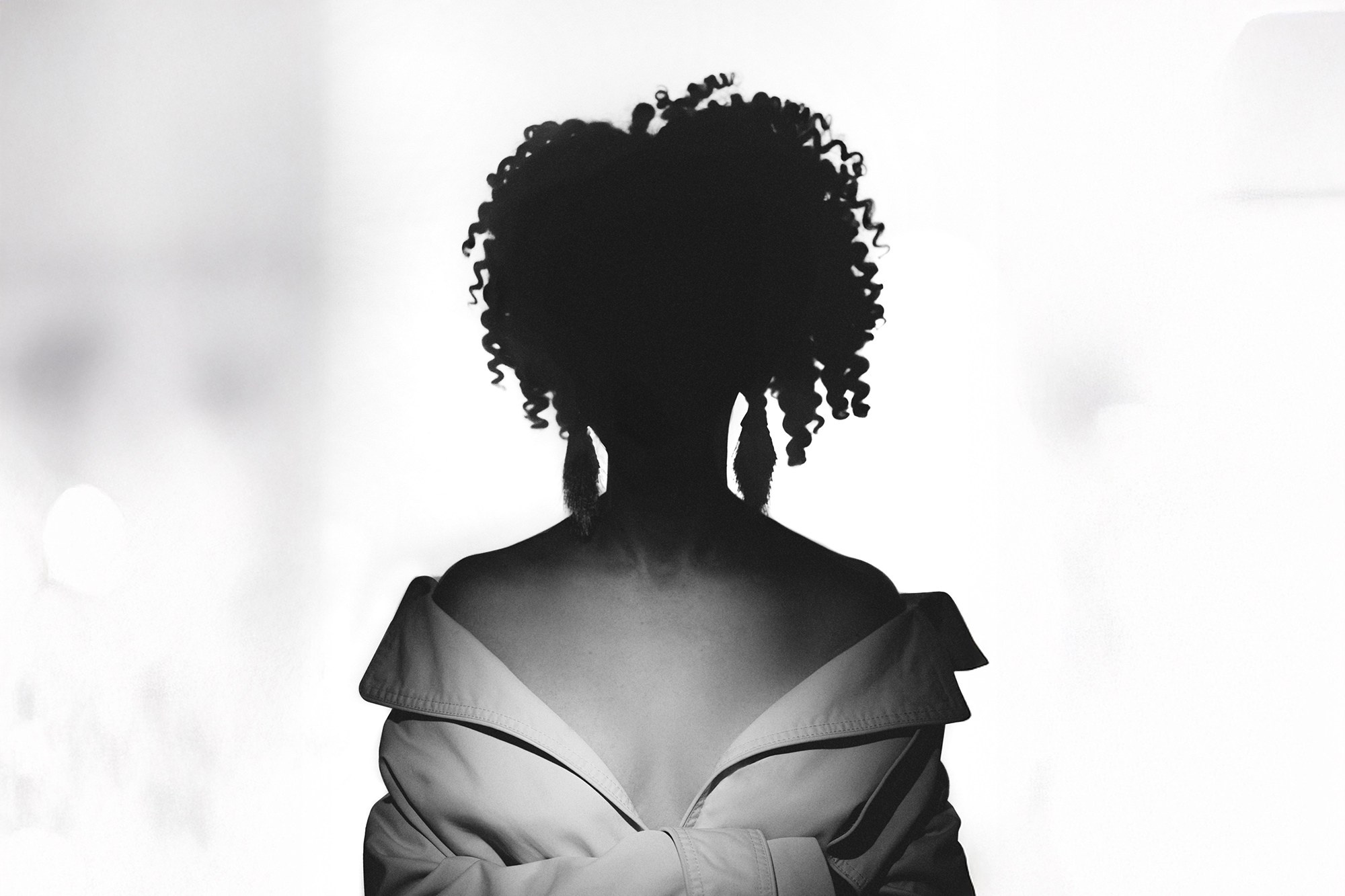 Графичный черно-белый силуэт женщины с пышными кудрявыми волосами и крупными серьгами. Контрастное освещение сзади (контурный свет) создает яркий ореол вокруг фигуры на светлом фоне. Портретный фотограф Галина Скорик.