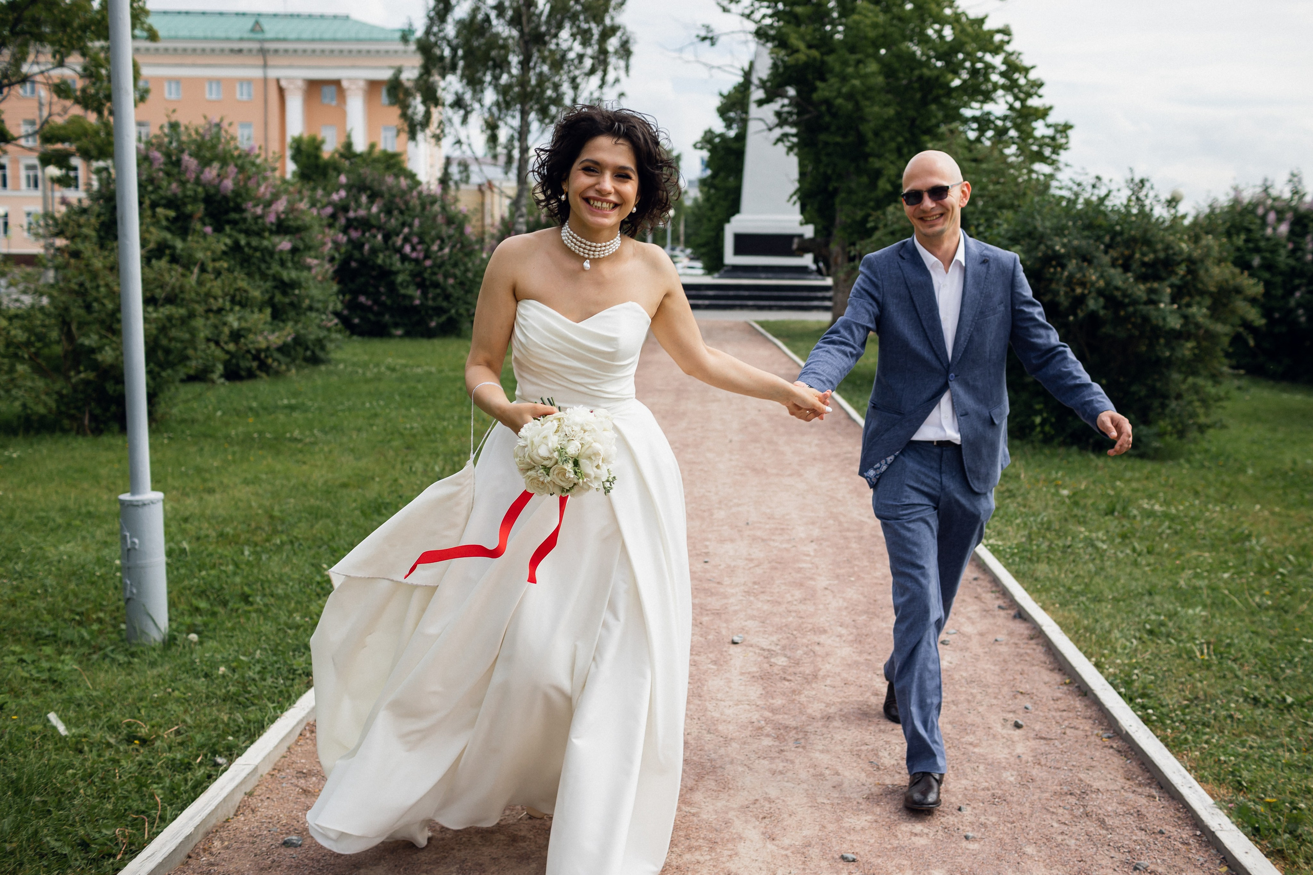 Счастливые жених и невеста бегут по аллее парка в Архангельске, улыбаясь камере на фоне цветущих клумб и красивых деревьев
