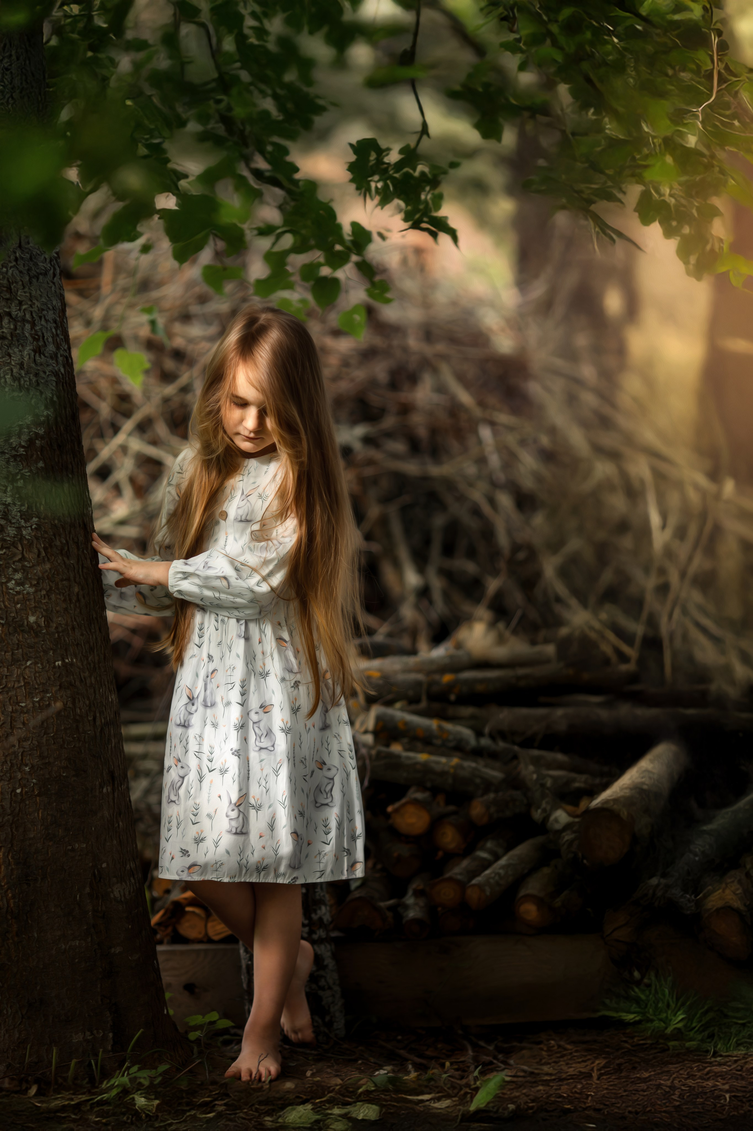 Девочка с длинными светлыми волосами в платье с зайцами стоит у дерева на фоне сложенных веток. Мягкий свет пробивается сквозь листву, создавая сказочную лесную атмосферу.