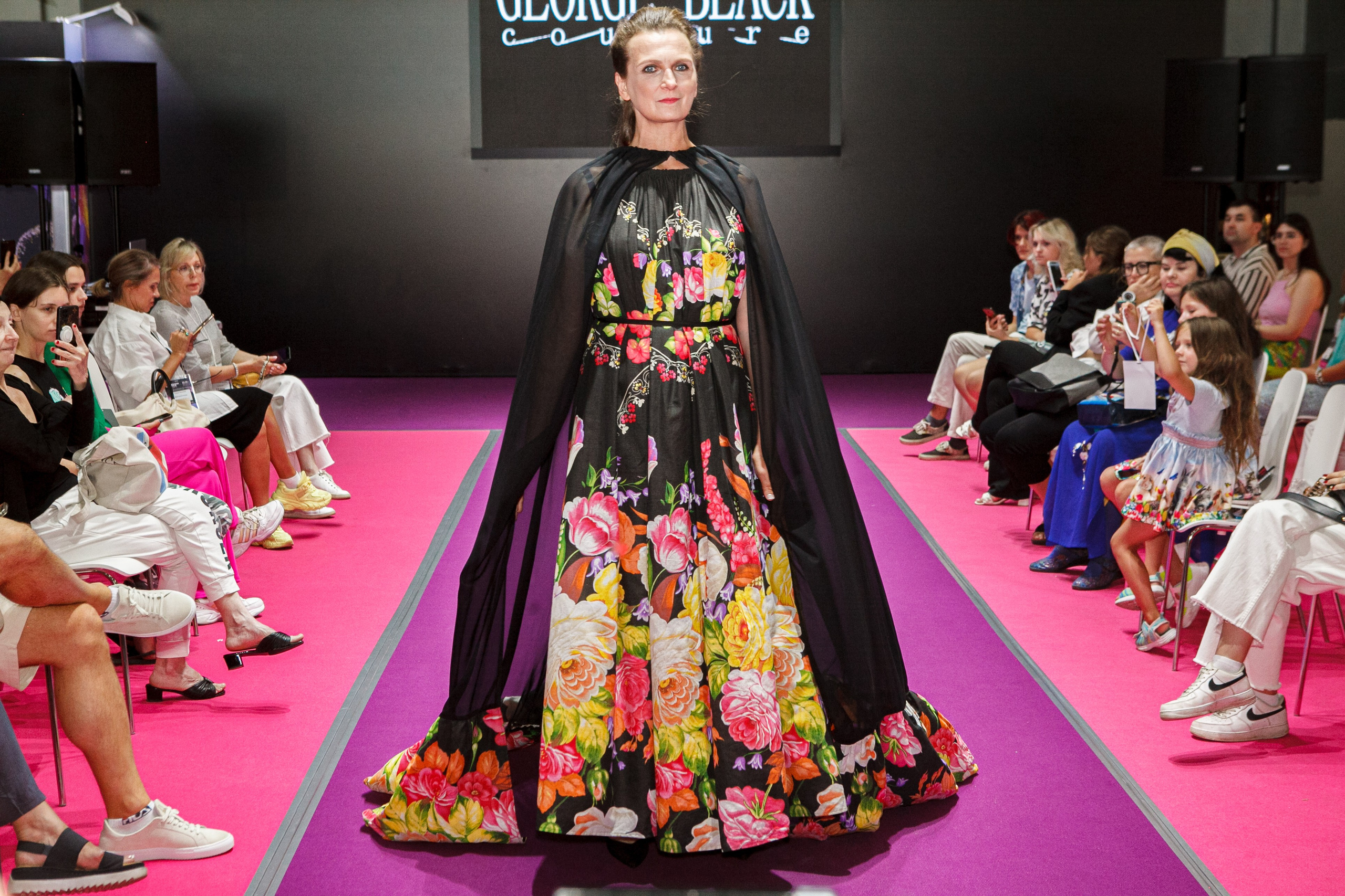  "Модели в ярких нарядах проходят по подиуму во время показа мод, освещенные яркими софитами и окруженные восторженными зрителями."