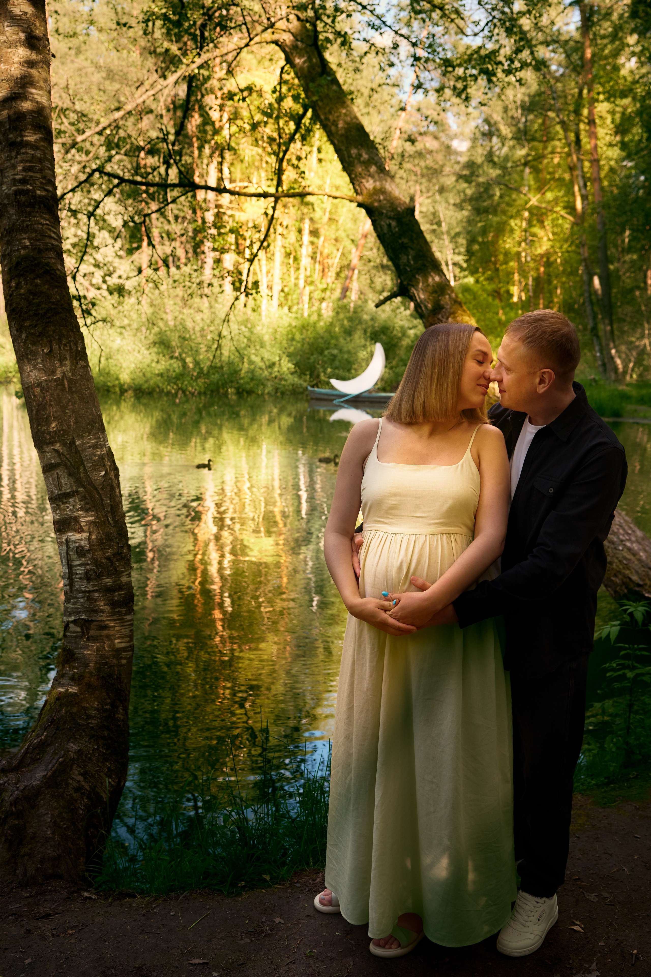 Фотография матери и ребенка в полный рост: пара стоит у озера на закате и обнимает беременную женщину, одетую в светлое платье.