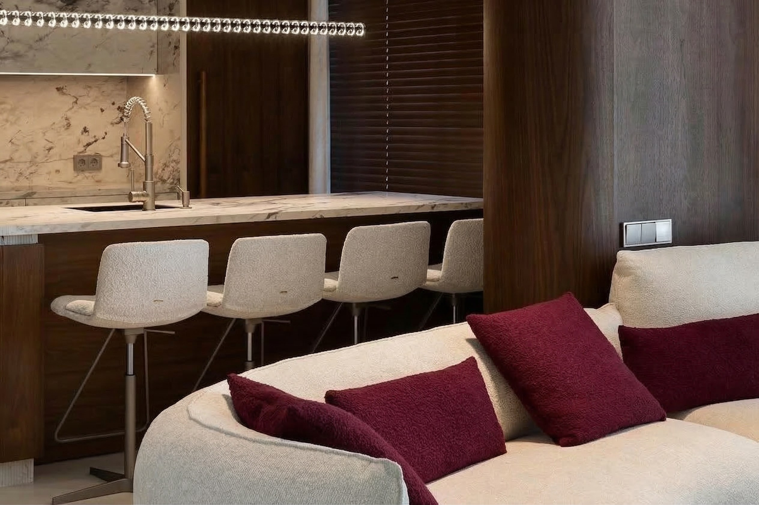 Деталь интерьера в ЖК ONYX Deluxe: светлый диван с бордовыми подушками на переднем плане, за ним — кухонный остров с мраморной столешницей и барными стульями. Дизайн Валерии Котович.