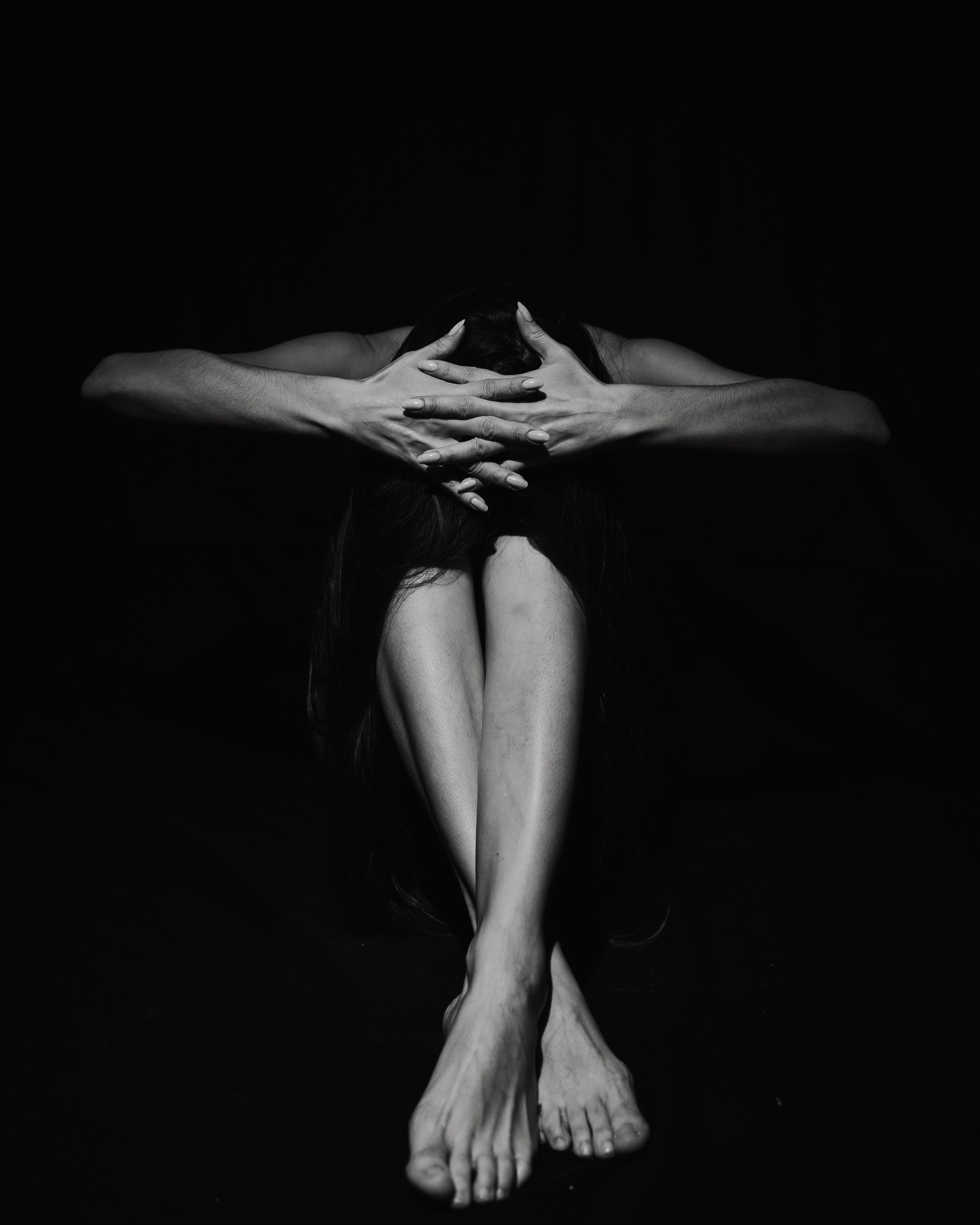 Изображена женщина, сидящая на полу, подтянув колени к груди и обхватив их руками. Ее голова опущена, волосы закрывают лицо. Похоже, что она испытывает сильную боль или дистресс. Изображение черно-белое, а фон темный, что усиливает ощущение изоляции и отчаяния.