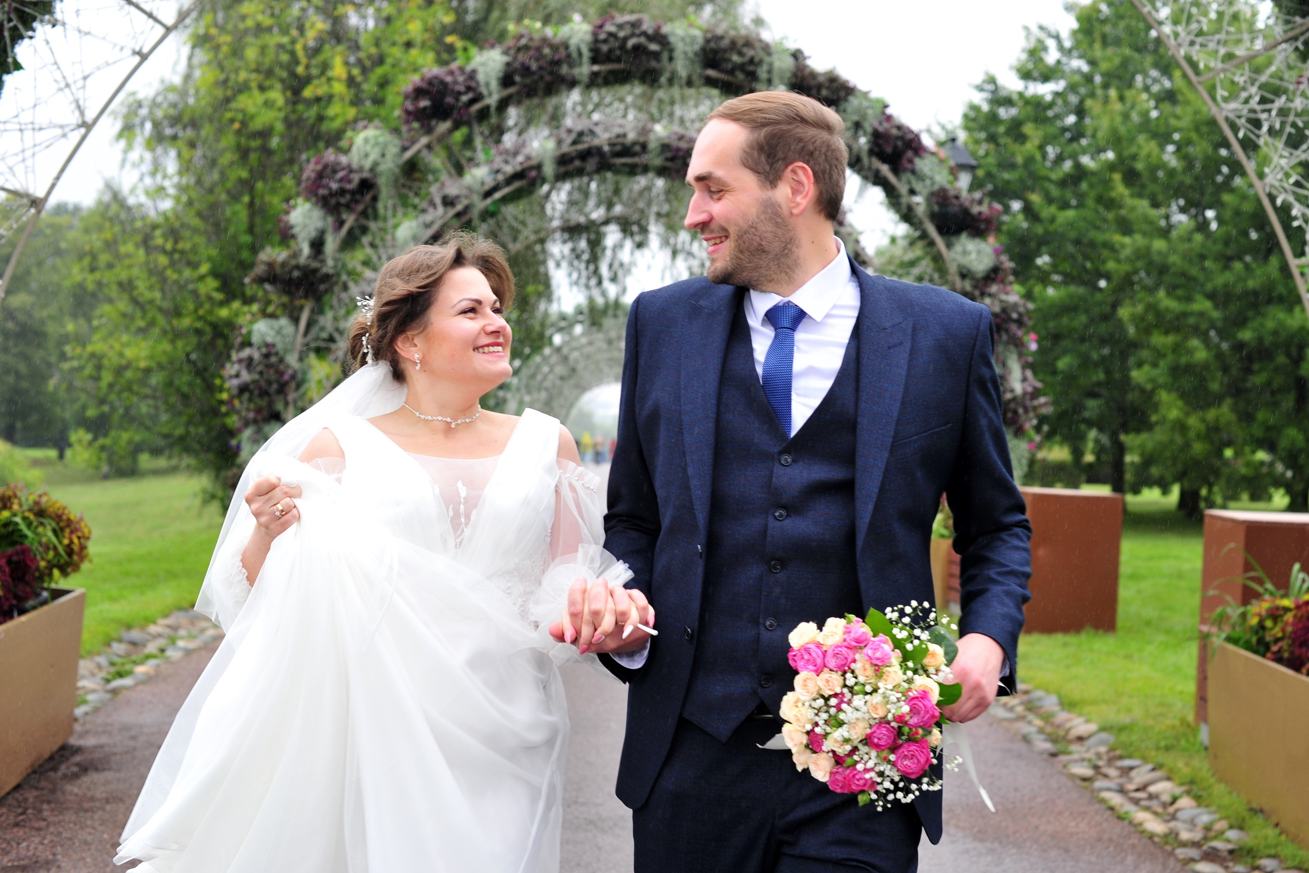 Свадьба в Царицыно пасмурным летним днем, новобрачные идут по влажной дорожке парка и держатся за руки на фоне зеленых арок из живых цветов 